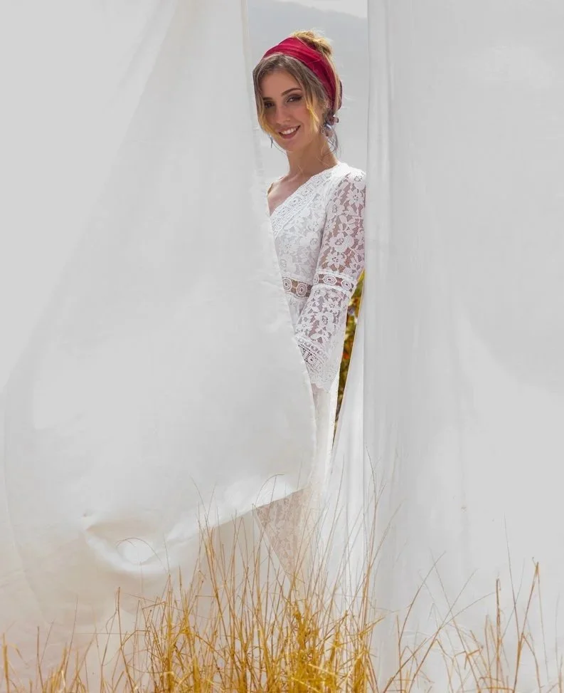 Femme souriante portant une robe blanche en dentelle, coiffée d'un bandeau rouge, regardant à travers une ouverture de tissu blanc avec des herbes jaunes au premier plan.