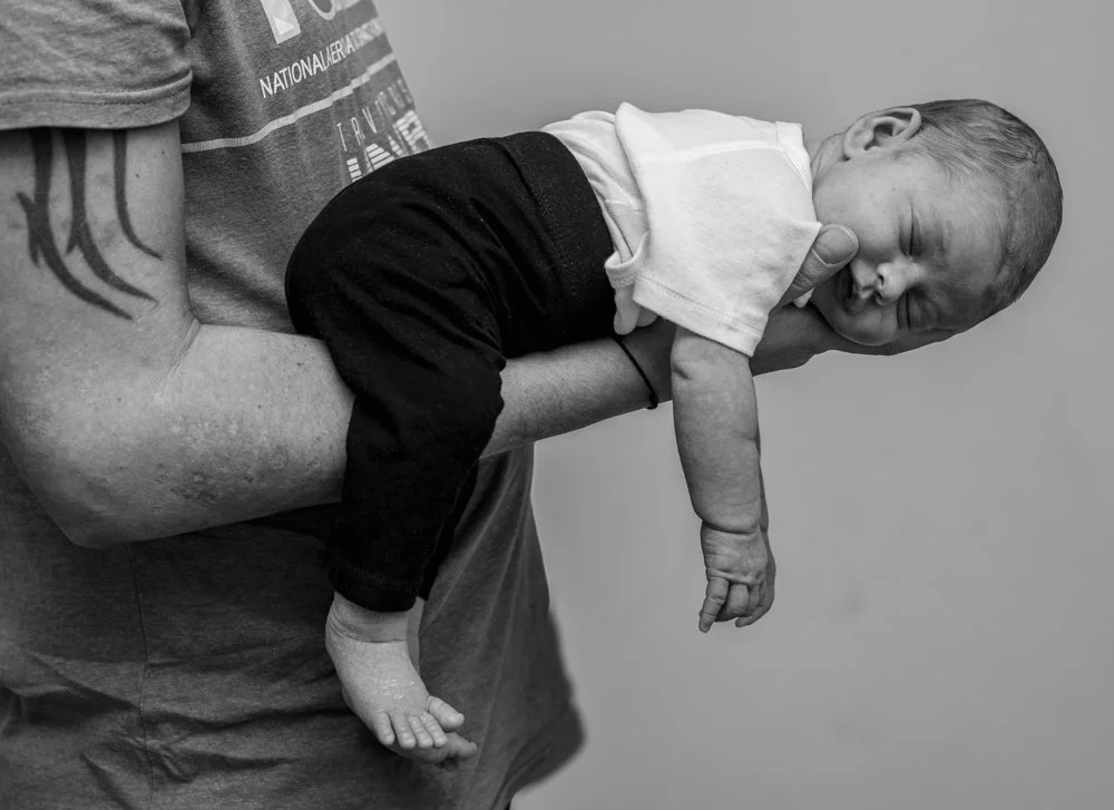 Un bébé allongé sur le bras d'une personne dont le visage n'est pas visible, en attente d'un sourire ou d'une réaction, en noir et blanc.