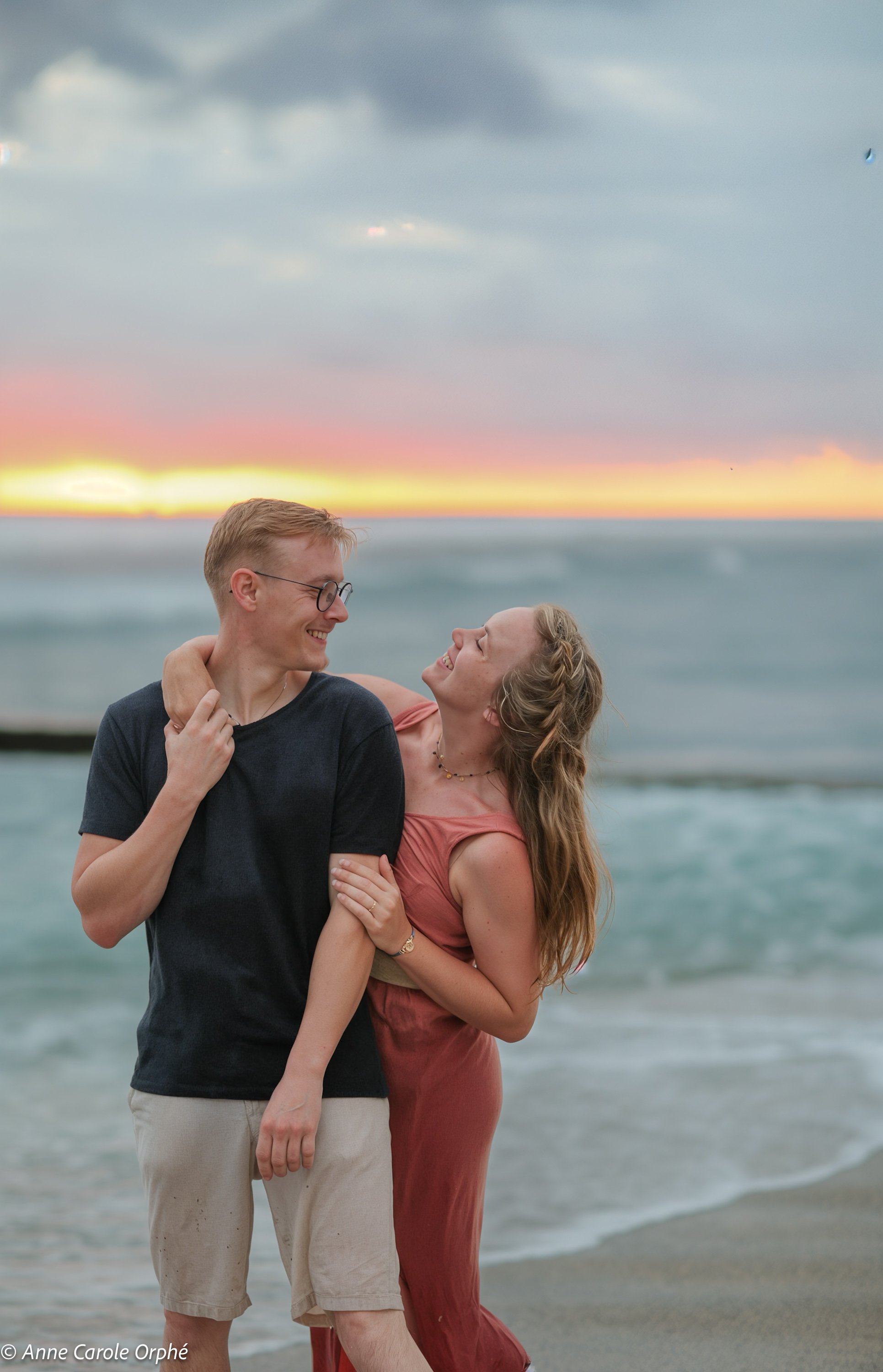 Un couple riant et se tenant par la taille sur la plage au coucher du soleil, avec la mer en arrière-plan et un ciel orageux.