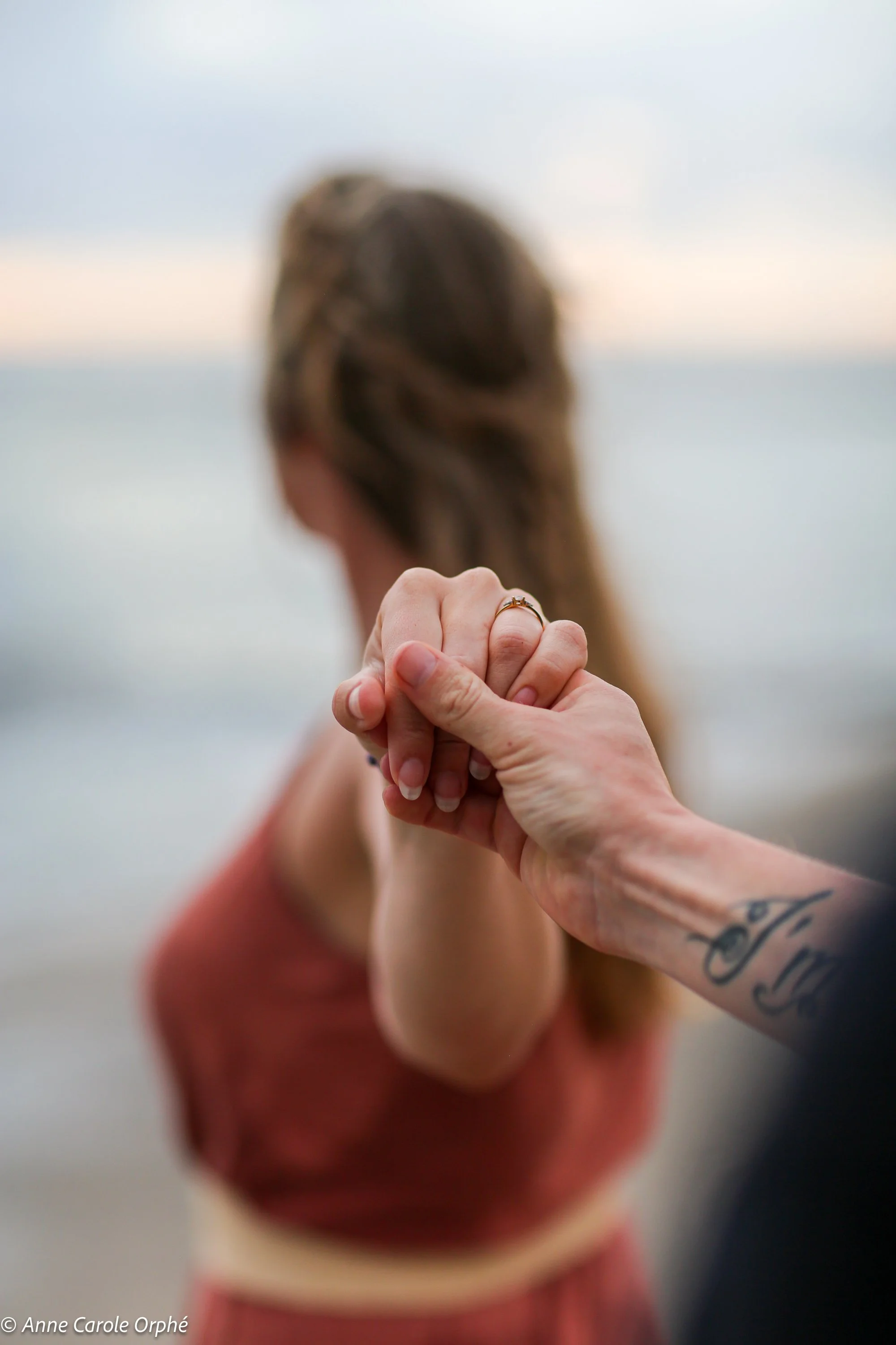 Une femme et une personne masculine se tiennent par la main, la femme porte une bague à l'annulaire et a un tatouage sur le bras. La femme a les cheveux bruns longs, la scène semble se dérouler près d'une étendue d'eau.