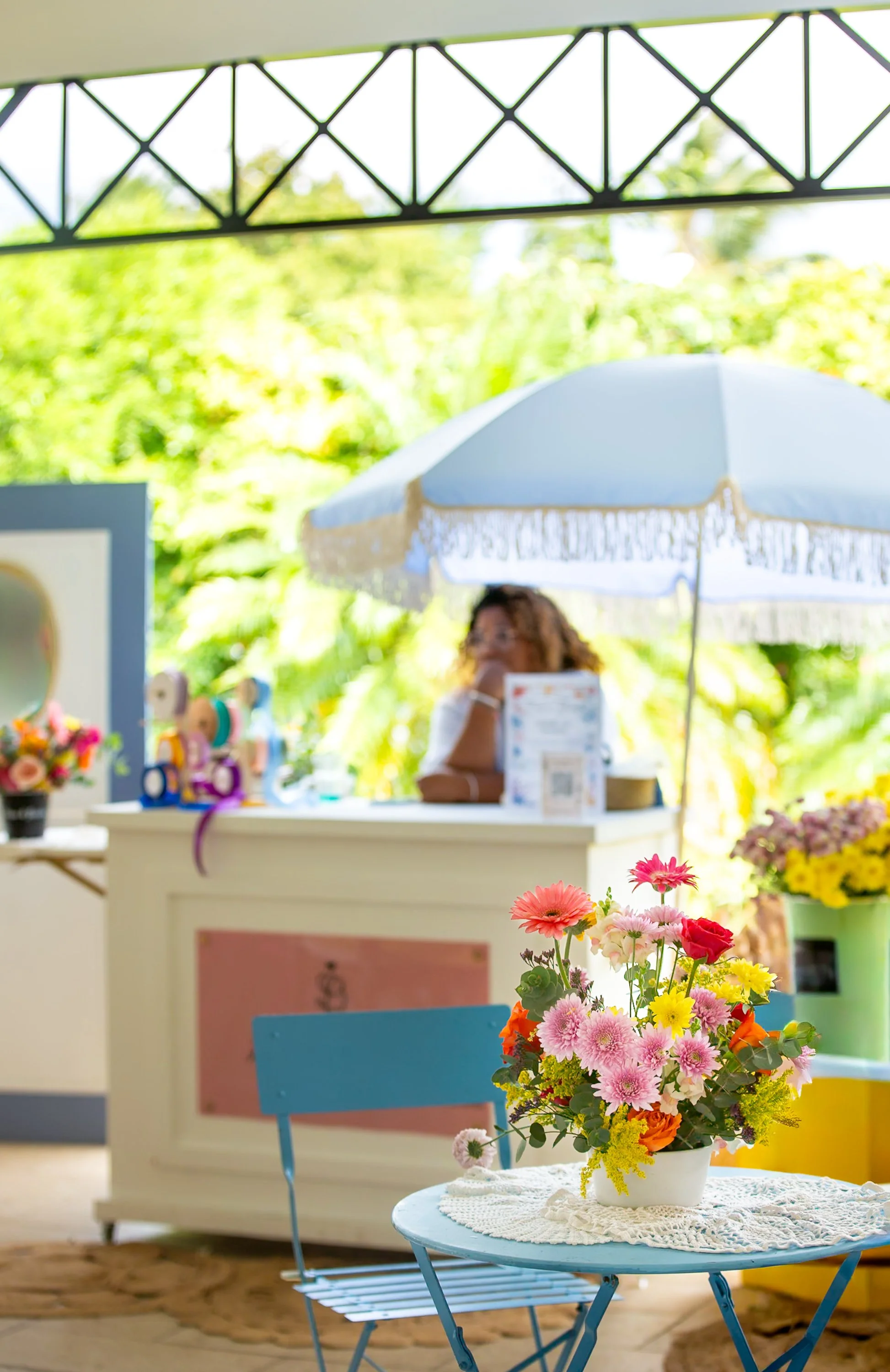Une table ronde bleue avec un bouquet de fleurs de différentes couleurs dans un vase blanc, et une chaise bleue à côté, dans un décor lumineux avec des fleurs en premier plan.