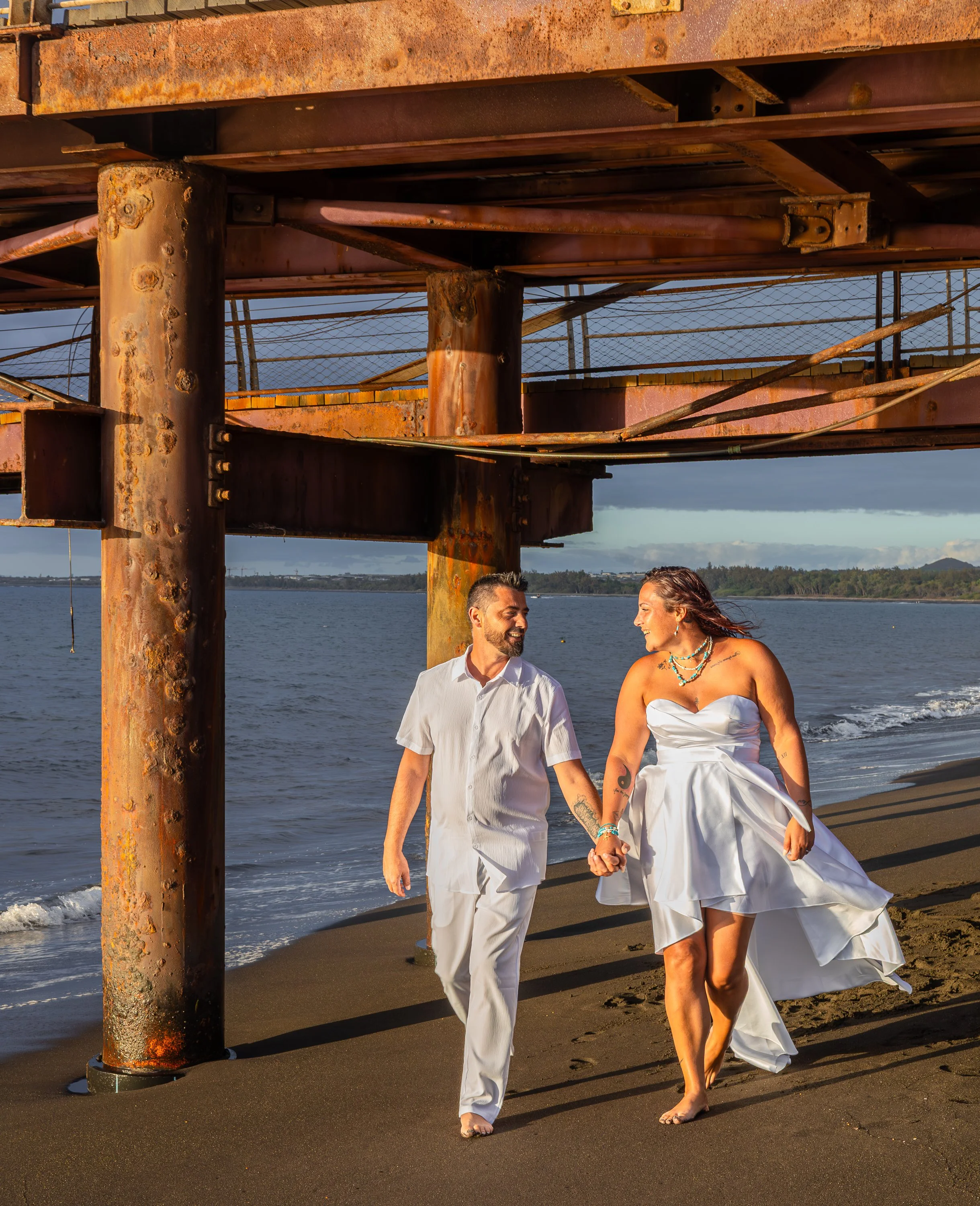 Un couple marchant main dans la main sur la plage, tenant la main, avec des bâtiments en arrière-plan, portant des vêtements blancs, à l'époque du coucher de soleil.