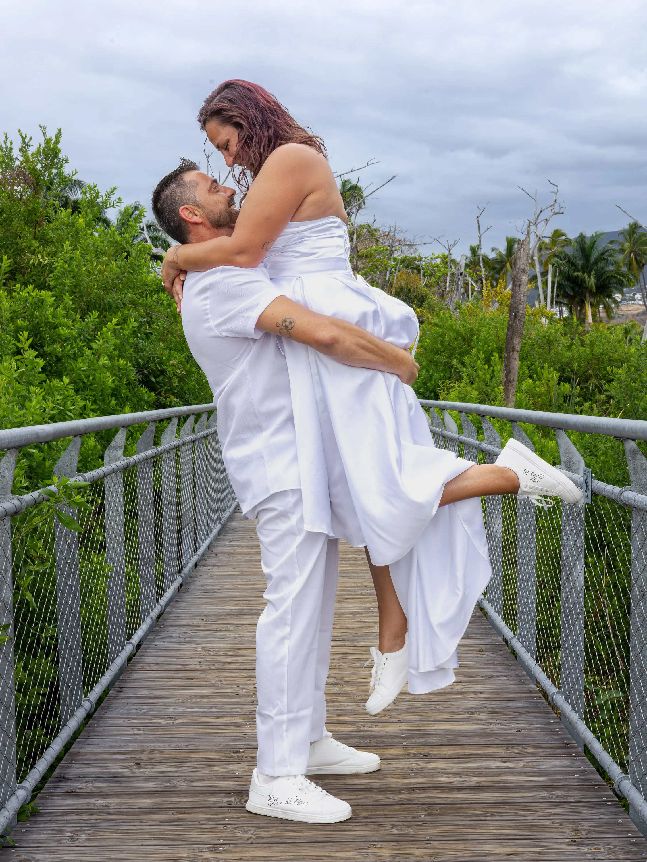 Un couple danse et sourit sur un pont en bois, entouré de verdure et de palmier, sous un ciel nuageux.