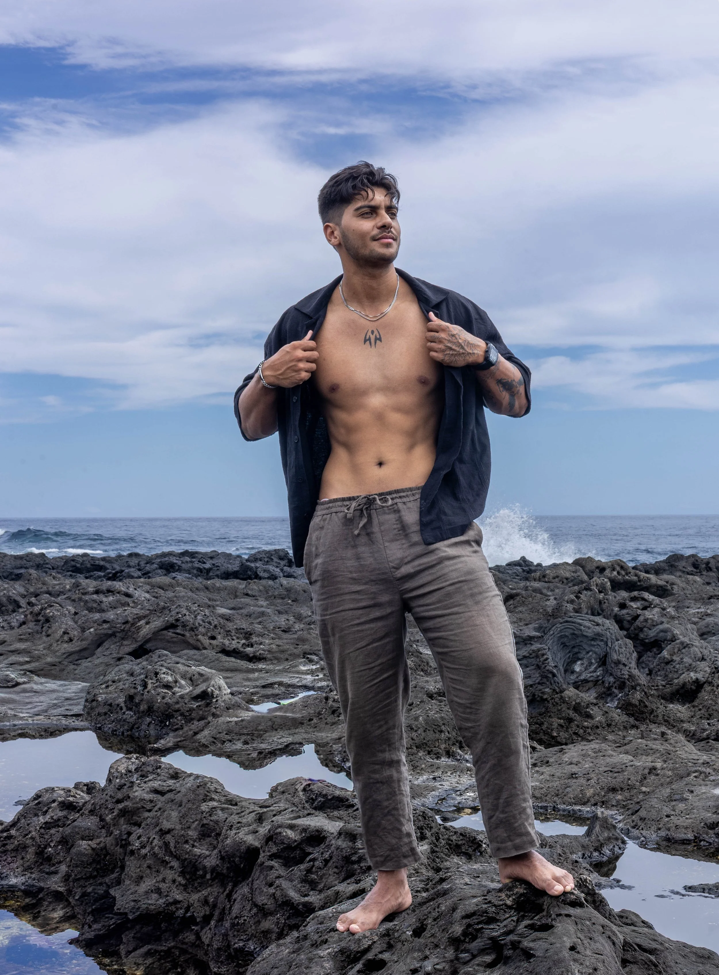 Un homme debout sur des rochers près de la mer, portant une chemise ouverte, montrant un tatouage sur la poitrine, avec un ciel nuageux en arrière-plan.