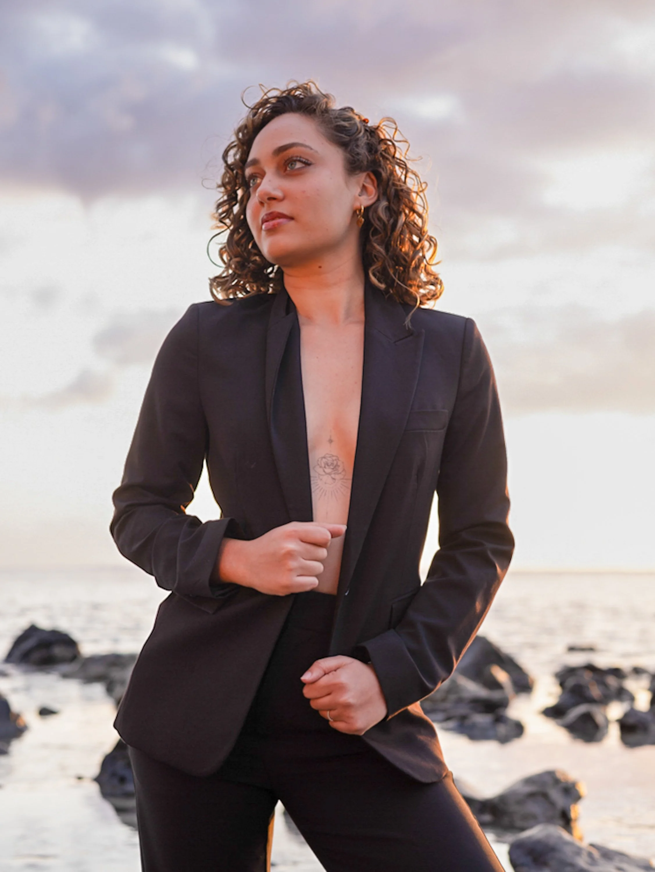 Une femme aux cheveux bouclés portant un tailleur noir se tient sur la plage devant la mer au coucher du soleil, regardant vers la gauche.