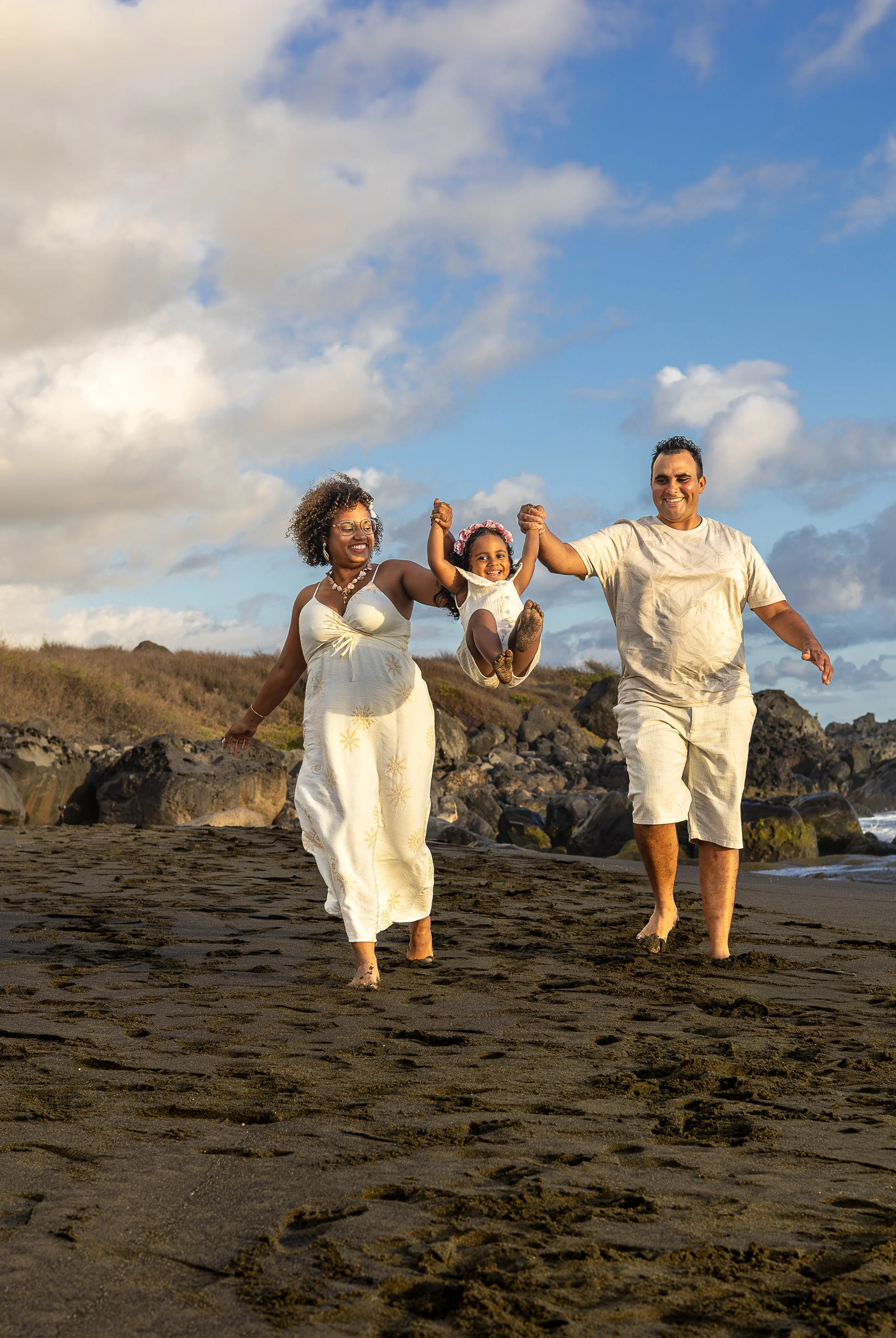 Un groupe de trois personnes, adultes et enfant, marchant sur la plage, rires, vêtements légers, paysage côtier avec rochers et ciel partiellement nuageux.