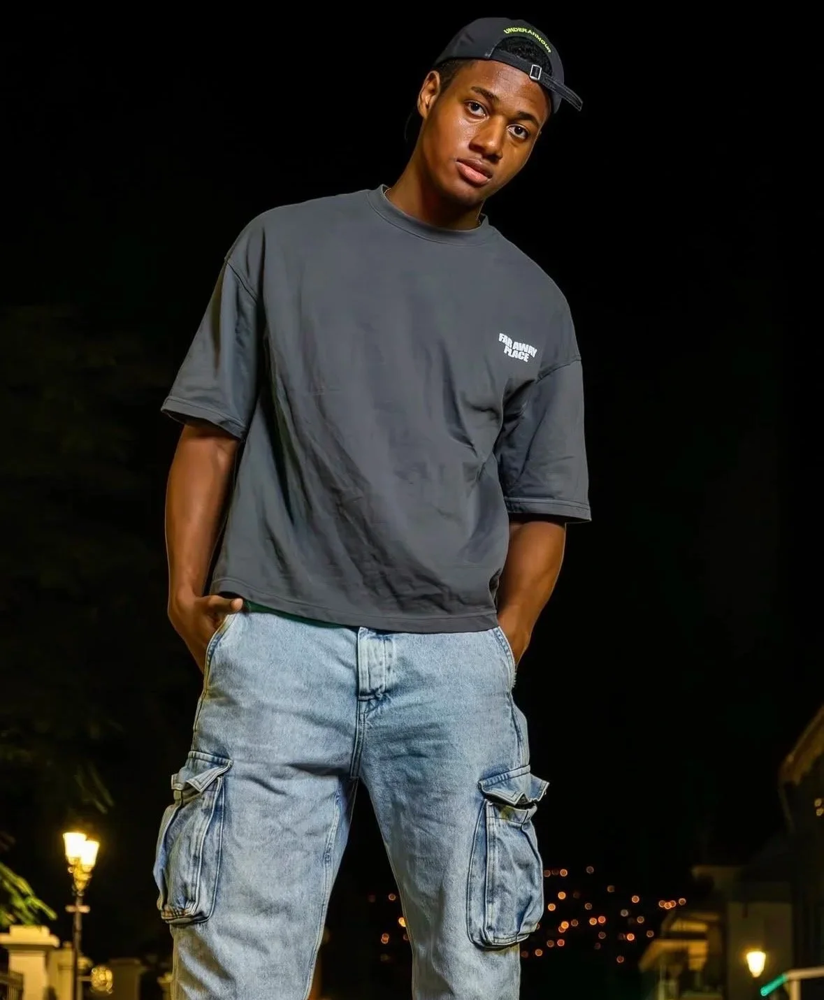 Jeune homme portant un t-shirt gris et un pantalon cargo, portant une casquette à l'envers, dans un cadre nocturne.