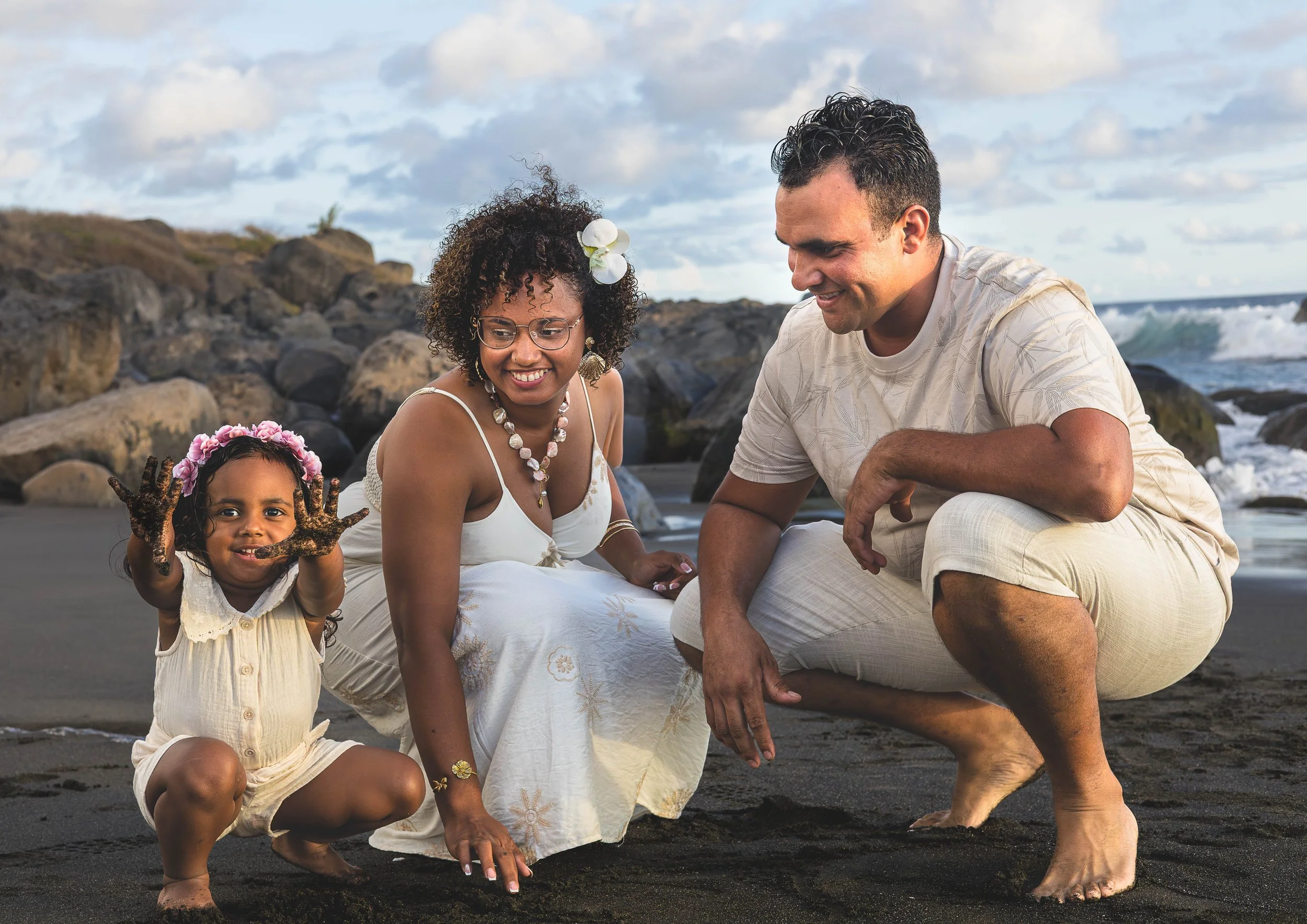 Famille sur la plage, accroupie dans le sable, avec des rochers en arrière-plan, en regardant vers le sol, souriant, lors d'une journée ensoleillée.