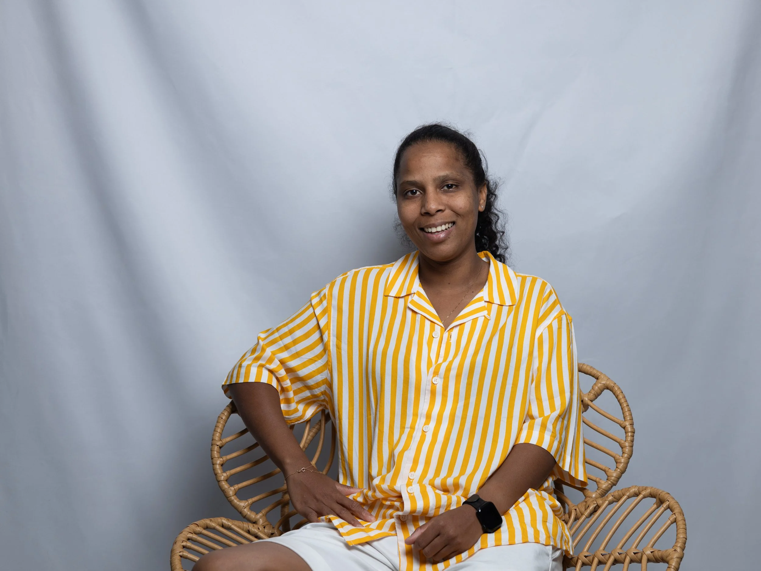 Une femme souriante portant une chemise rayée jaune et blanche, assise sur un fauteuil en osier.