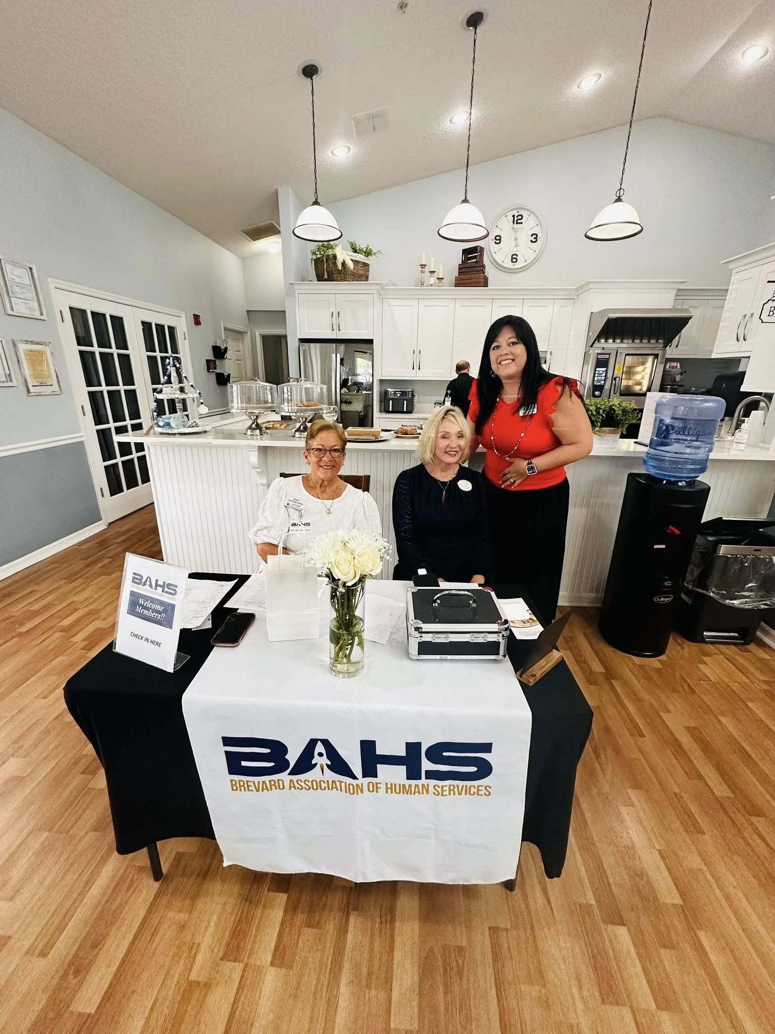 Three women at a Brevard Association of Human Services (BAHS) table, with two sitting and one standing, in a well-lit room with kitchen appliances and decorations in the background.