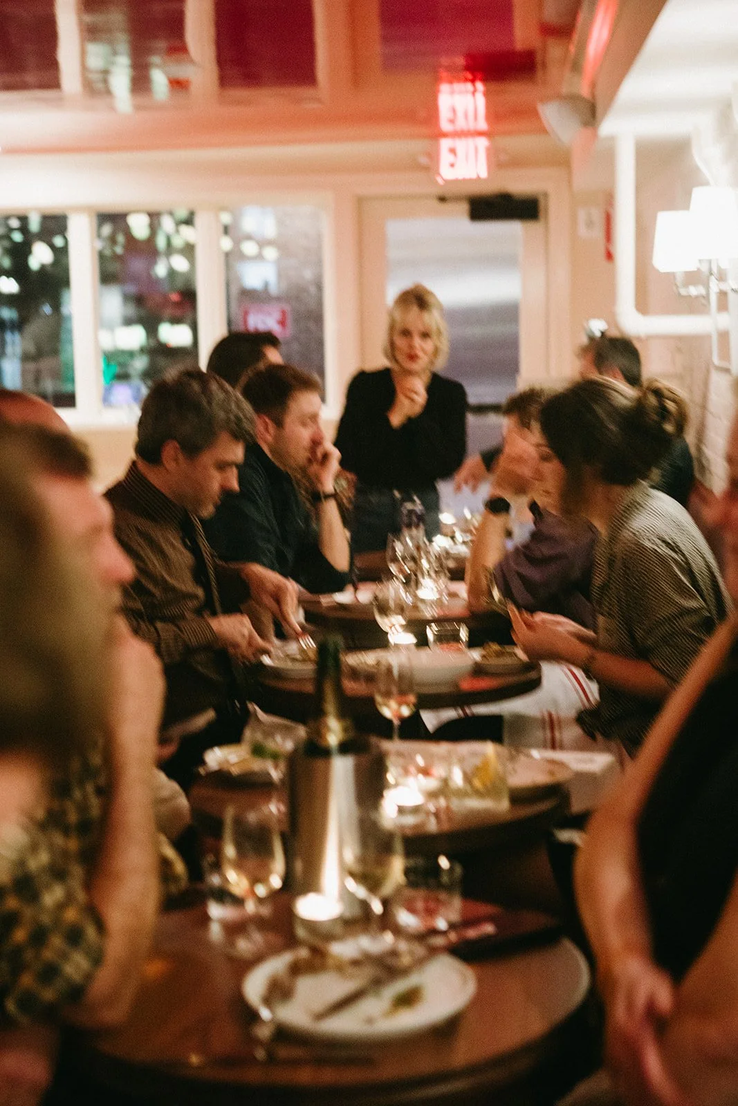 A group of people dining together at a restaurant with a woman standing, possibly explaining or addressing the group, illuminated with warm lighting, with wine glasses and plates on the table.