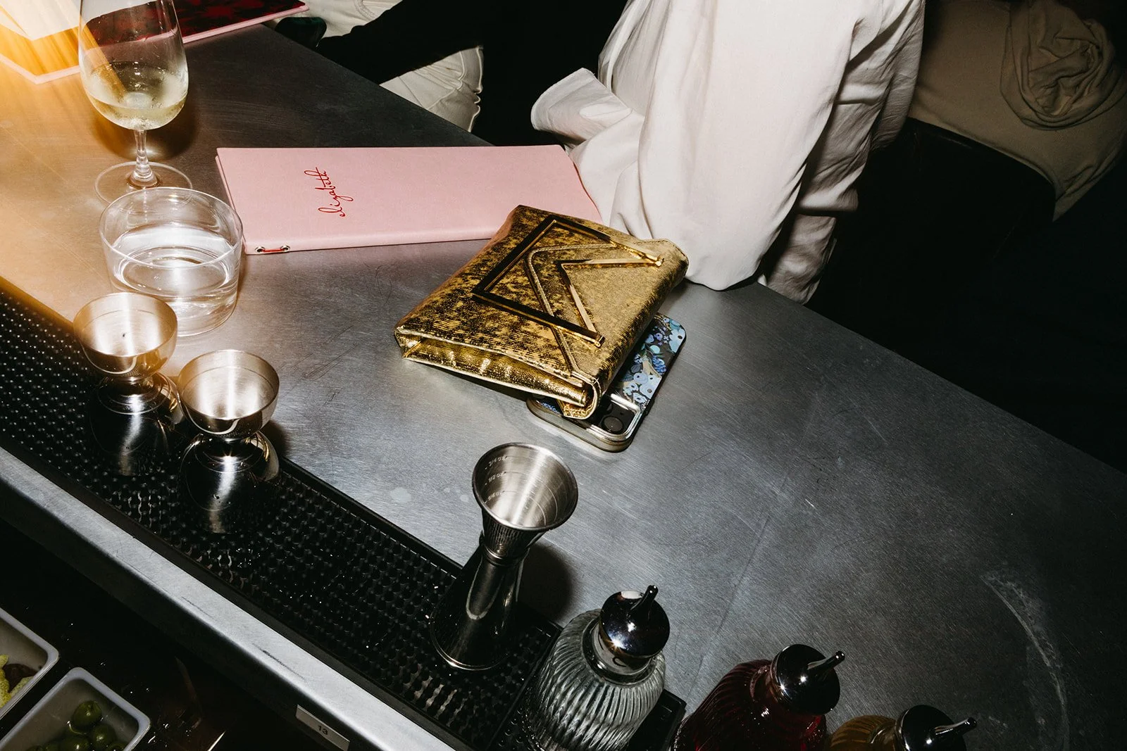 A bar counter with three metal shot glasses, a tall glass of wine, a glass of water, a pink menu, a gold and black patterned clutch, a smartphone with a blue floral case, and a person's arm in a white shirt resting on the counter.