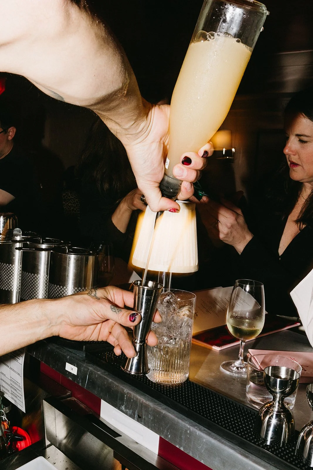 Bartender pours a yellow cocktail into a metal jigger at a bar, with patrons watching and drinks on the bar counter.