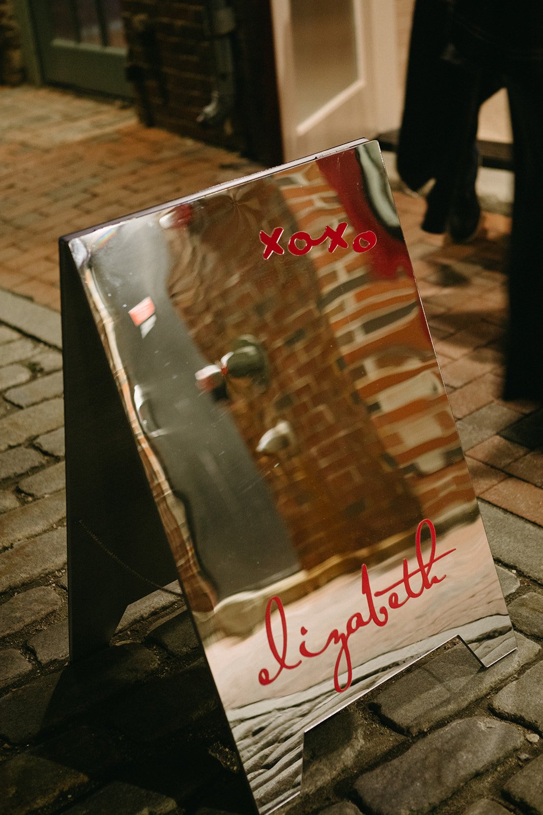 A reflective mirror sign on a cobblestone sidewalk with red cursive text that says "elizabeth" and red "xoxo" at the top, reflecting brick walls and partial figures of people.