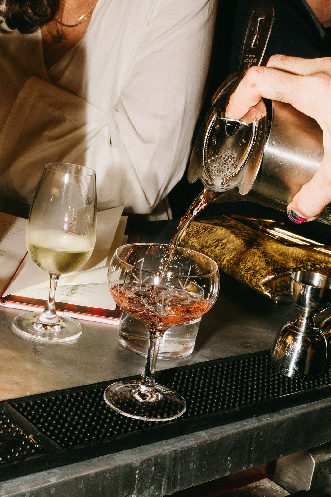 A bartender pours a pink cocktail into a glass, with a glass of white wine next to it on the bar counter.
