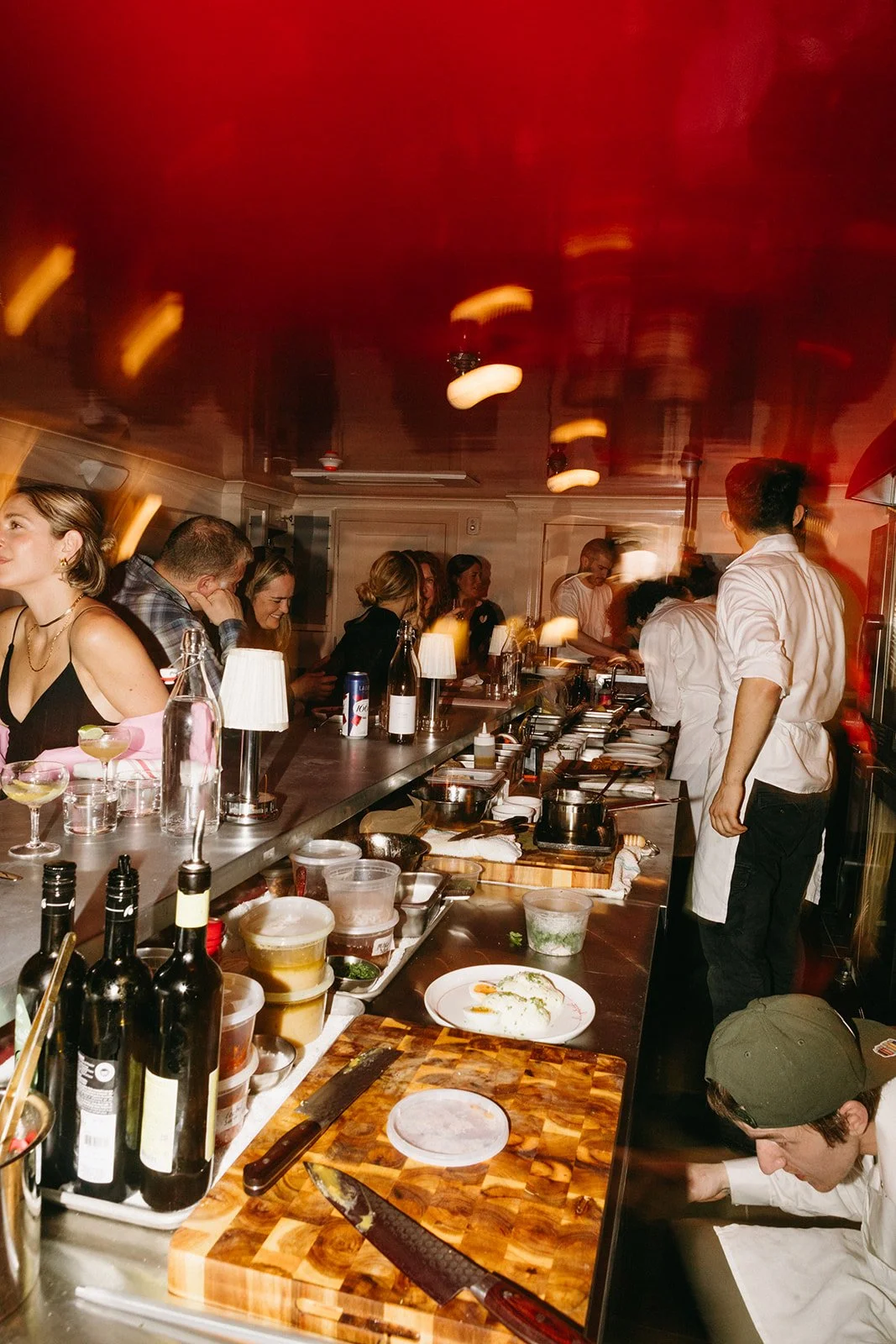 A lively restaurant kitchen with chefs preparing food behind a busy counter, and customers seated at the bar enjoying their meals, under warm lighting with a red ceiling.