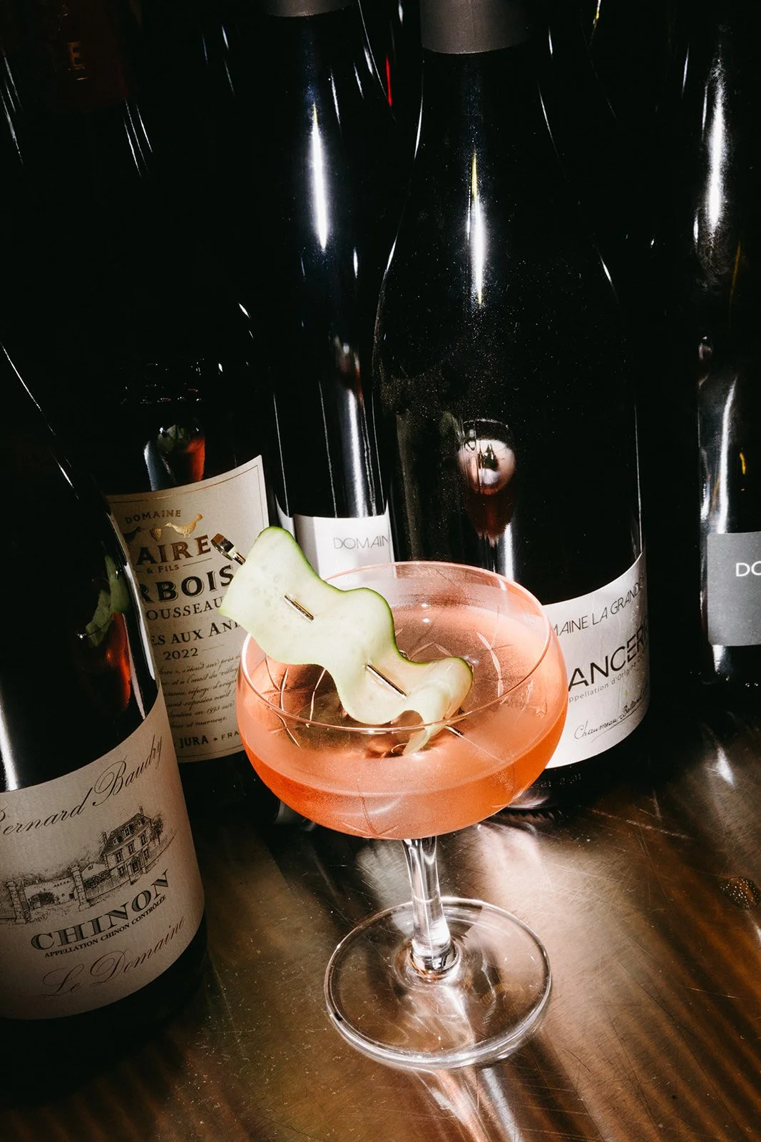 A pink cocktail garnished with a decorative cucumber strip in a coupe glass, surrounded by several bottles of red wine on a metallic surface.