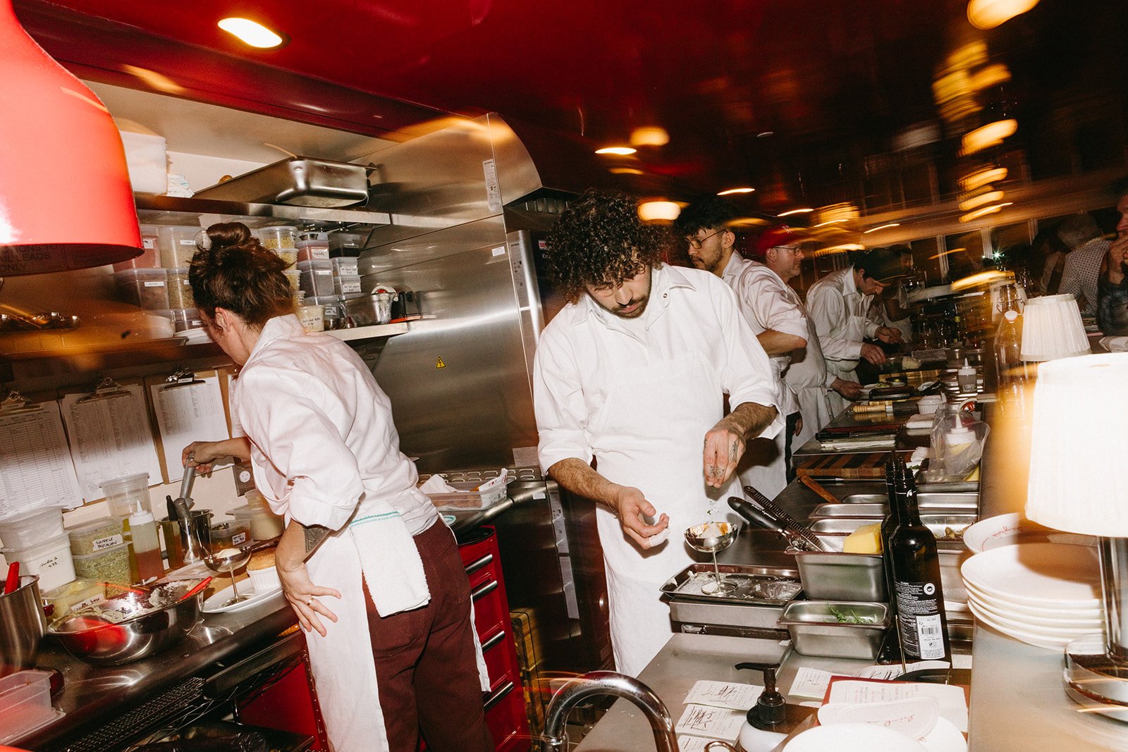 A group of chefs, dressed in white, working in a busy kitchen with stainless steel appliances, containers of ingredients, and various cooking tools.