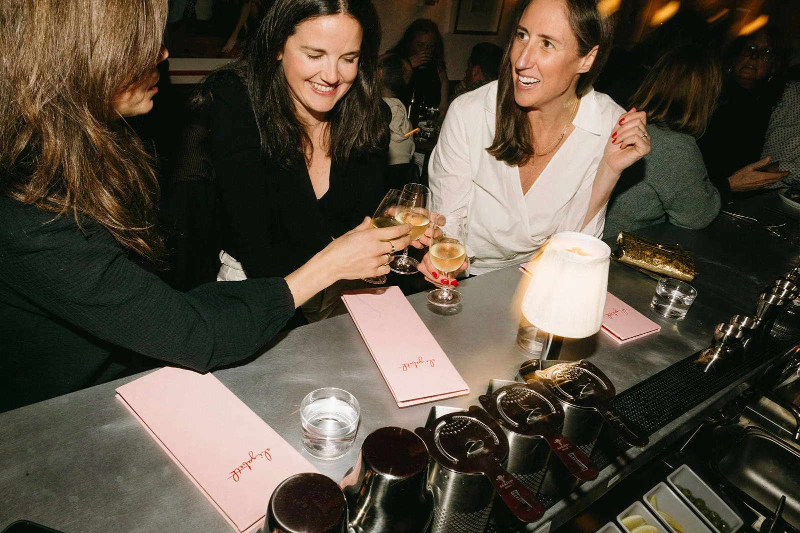 Four women sitting at a bar, clinking glasses of white wine, smiling and chatting.