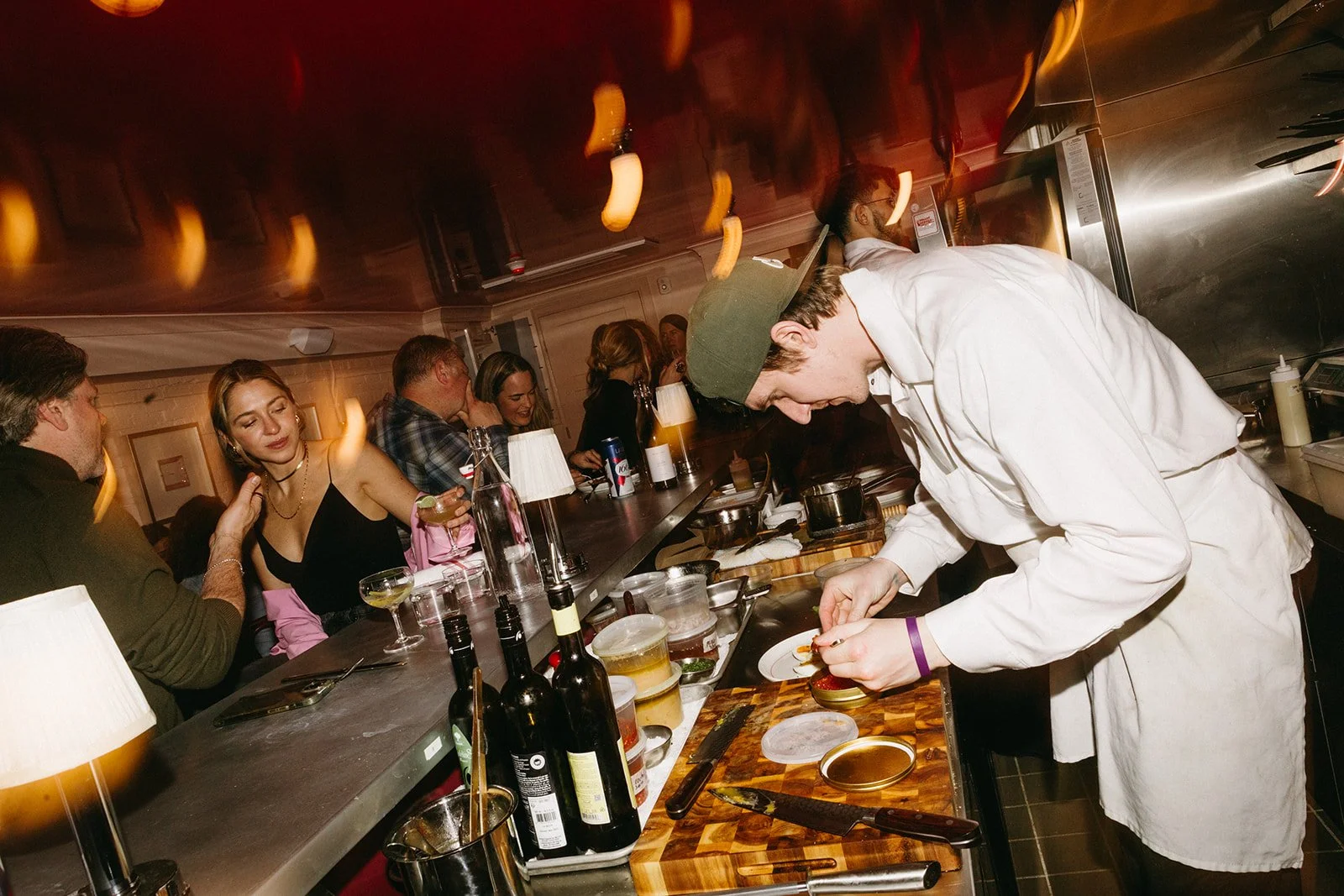 Chef plating a dish behind a bar full of patrons.