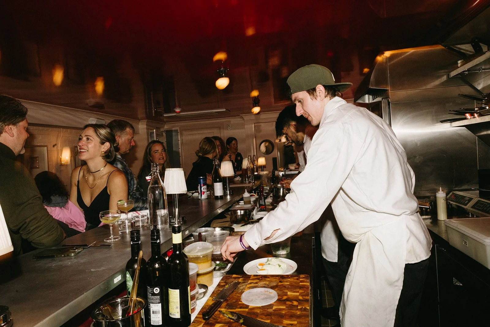 A chef in a white coat and green cap prepares food behind a bar counter while patrons enjoy drinks and conversation in a dimly lit restaurant.