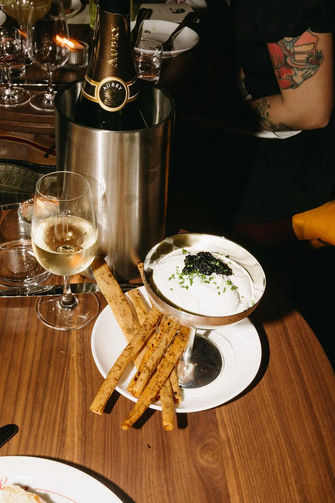 A bottle of champagne in a metallic ice bucket, a glass of white wine, a plate with bread sticks, and a bowl of creamy dip topped with black caviar and green herbs on a wooden table.