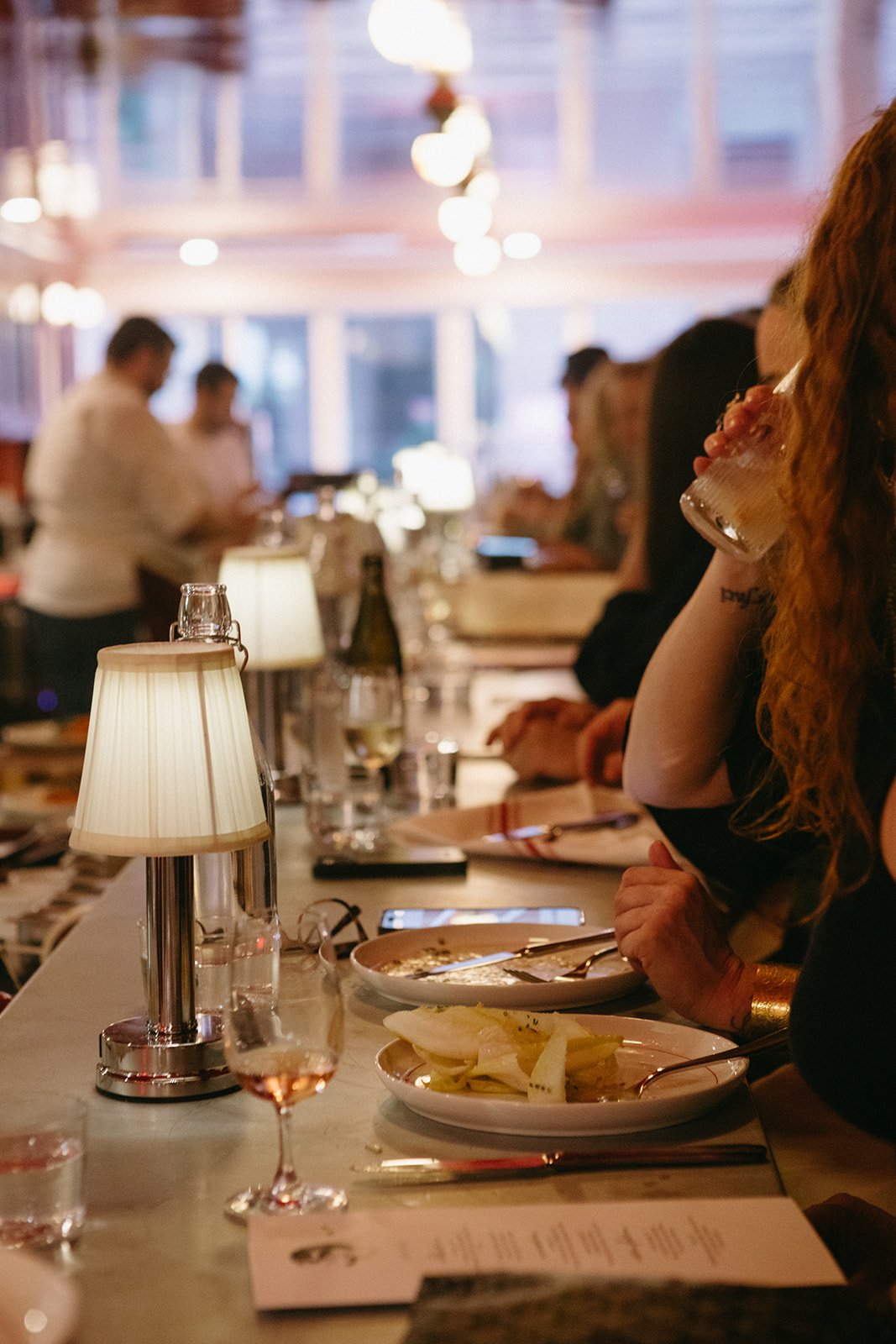 People dining at a restaurant with a bar, eating and drinking, with tables, glasses, lamps, and a blurred background.