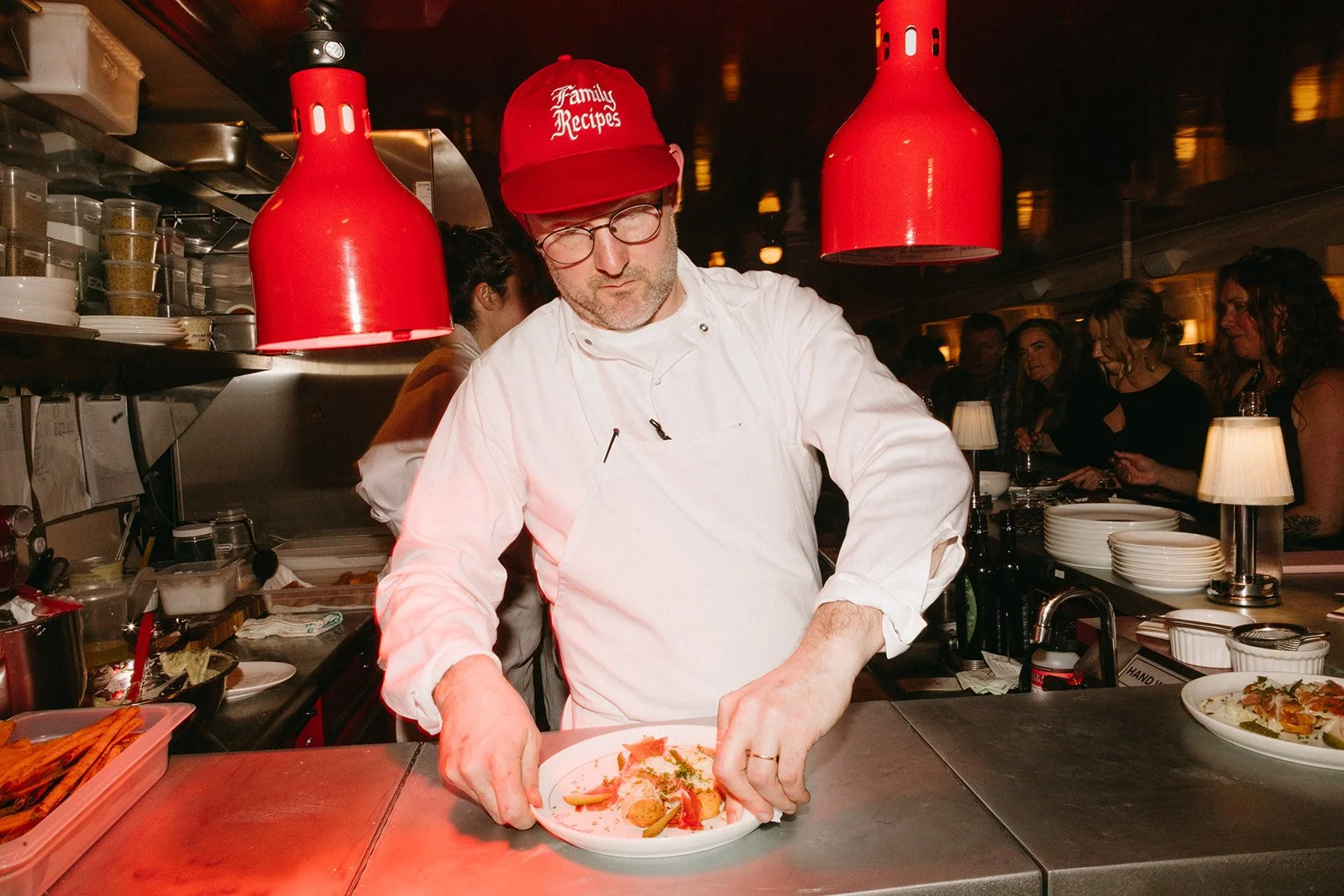 A chef wearing a red hat and glasses is plating a dish in a restaurant kitchen, with patrons dining in the background.