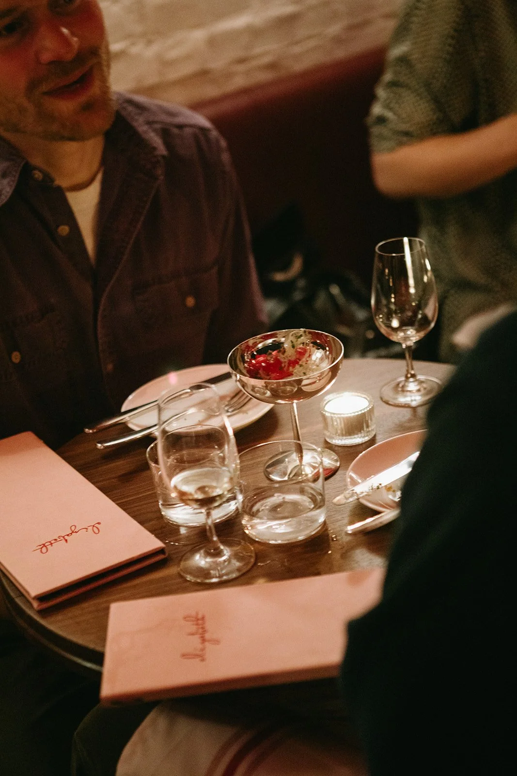 People seated at a round table with pink menus, glasses of water, a wine glass with a red cocktail, and a lit candle in a cozy, dimly lit restaurant.