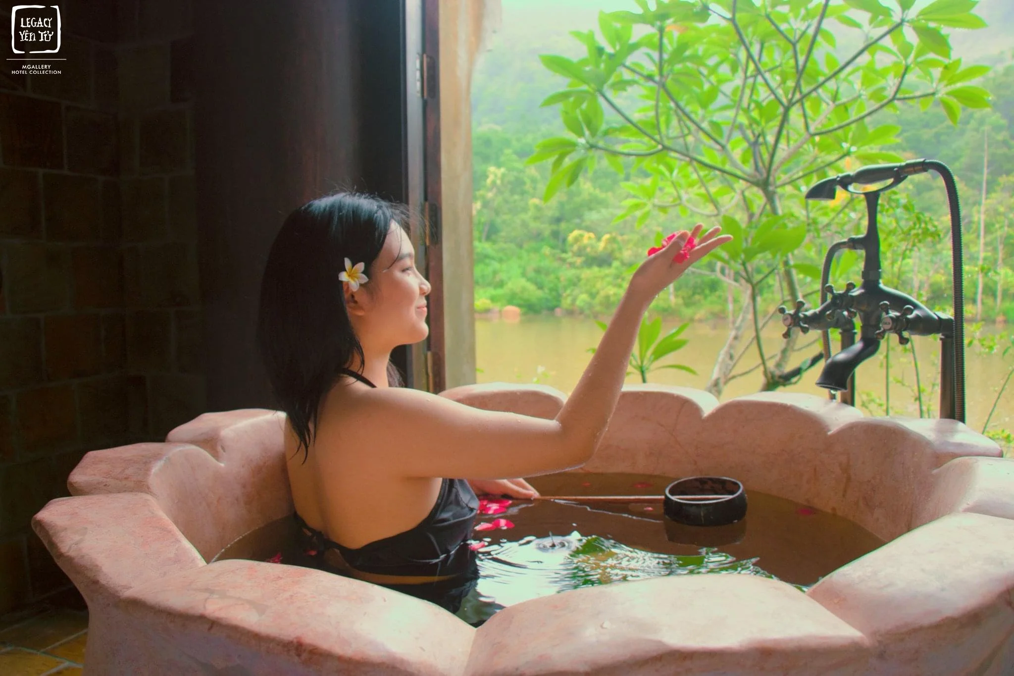 A woman with a white flower in her hair relaxing in a pink bathtub filled with water outdoors by a window, with greenery and trees visible outside.
