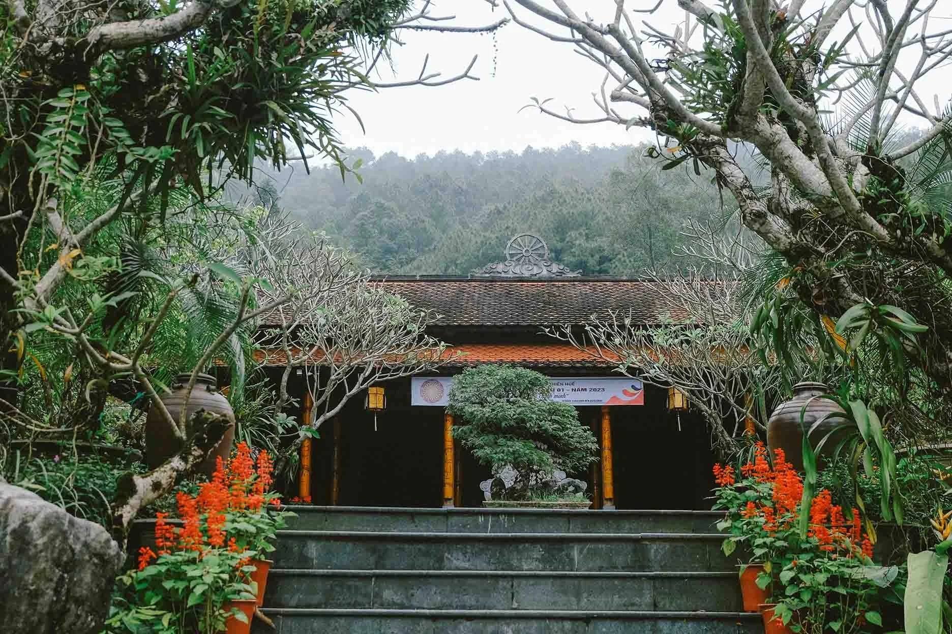 A traditional Vietnamese temple surrounded by lush greenery and trees, with potted plants and orange flowers in the foreground, and mountains in the background.