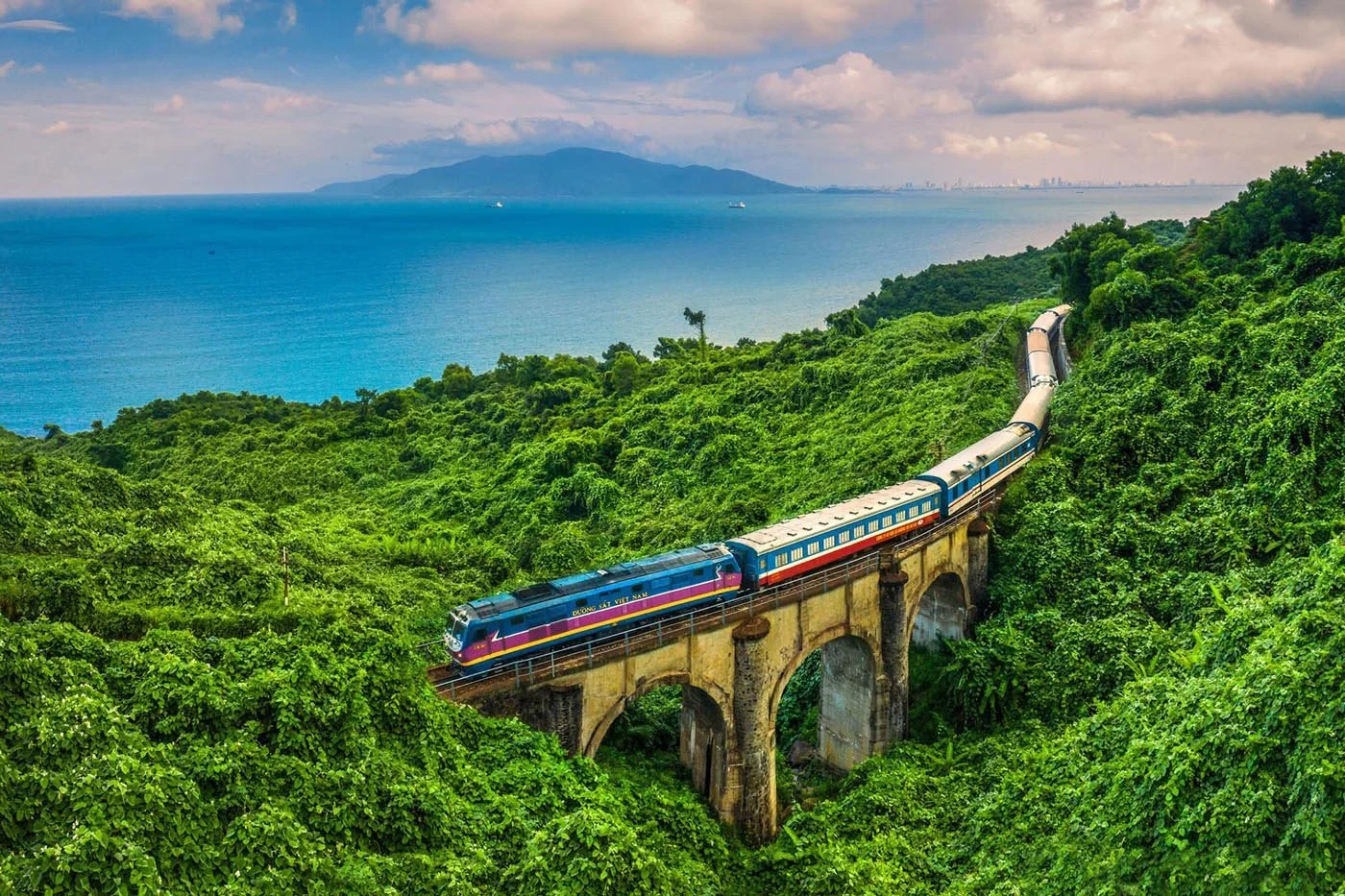 A train traveling on a bridge through lush green hills with a body of water and mountains in the background.