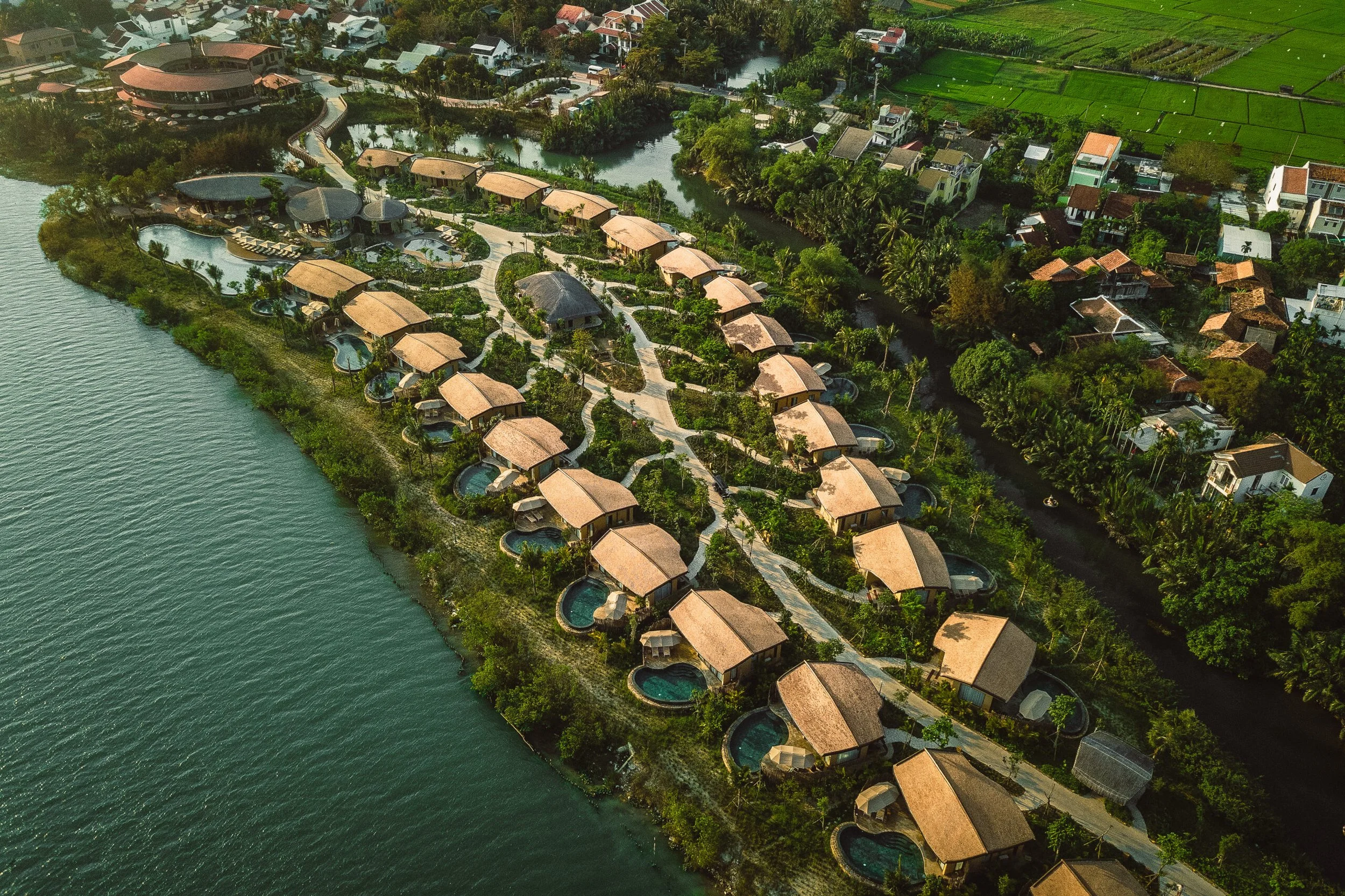 Aerial view of a tropical resort with overwater bungalows along a shoreline, surrounded by lush greenery, neighboring houses, and a body of water.