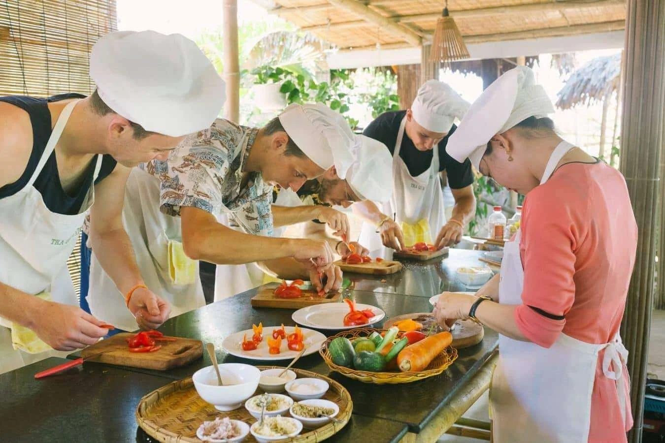 People cooking and preparing vegetables in a kitchen, wearing aprons and chef hats, with various ingredients on the table.