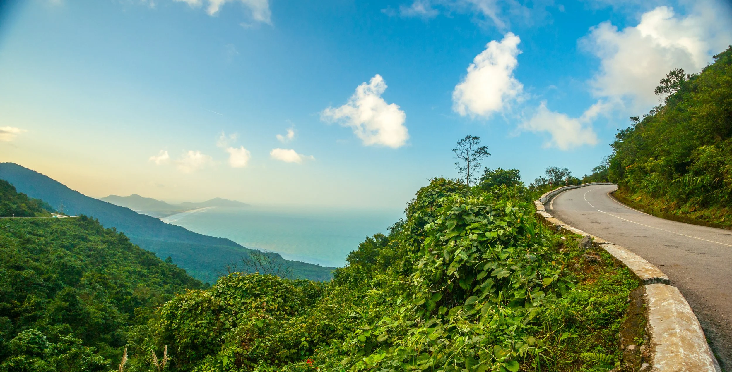 Winding mountain road surrounded by lush green vegetation with a view of a mountain range and the ocean in the distance, under a partly cloudy sky.