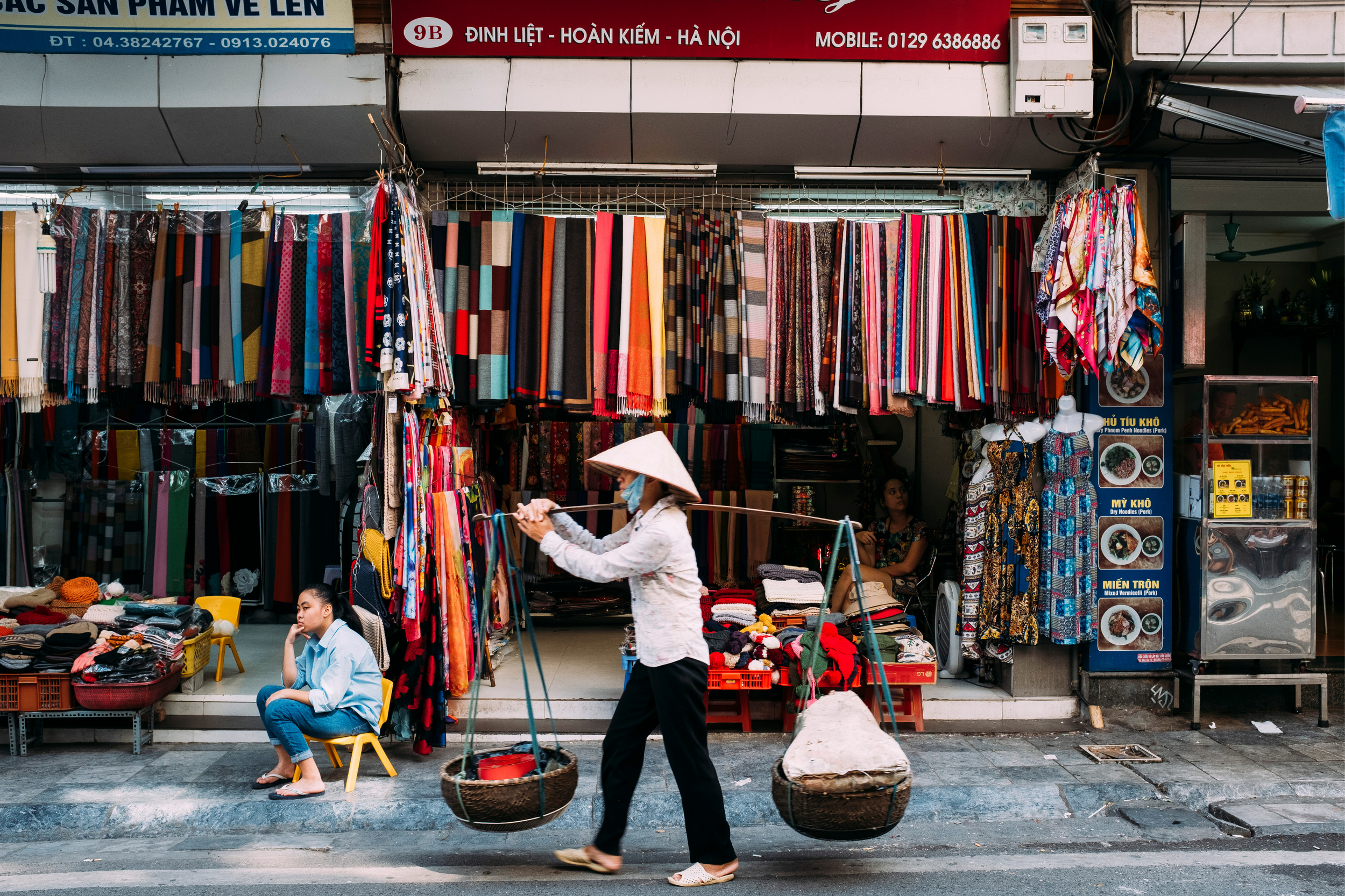 Street market scene with colorful fabrics hanging outside a shop, a woman wearing a traditional conical hat and face mask carrying baskets, a seated woman with a child nearby, and a sign with Vietnamese text.