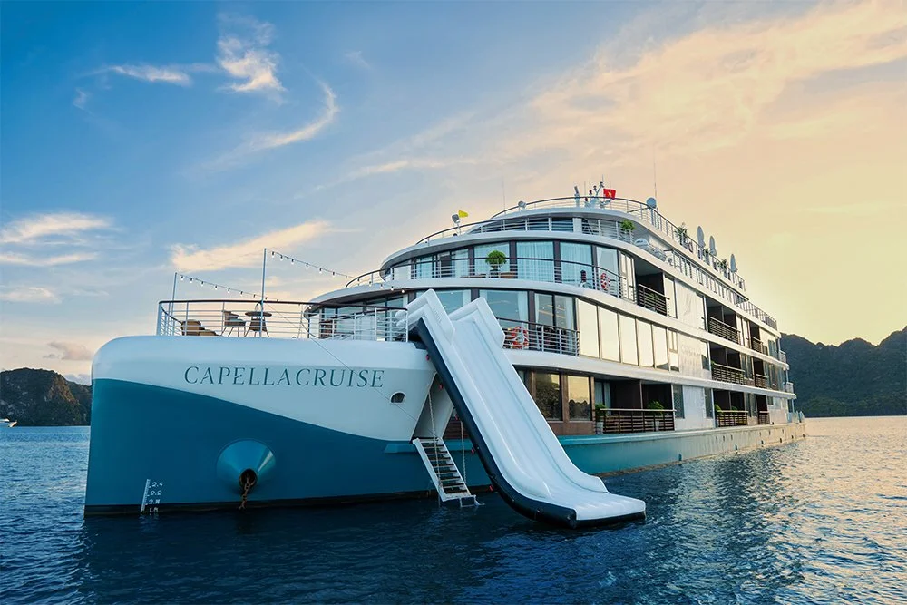 A large white cruise ship named Capella Cruise on calm water during sunset, with a water slide on the side and a mountainous landscape in the background.