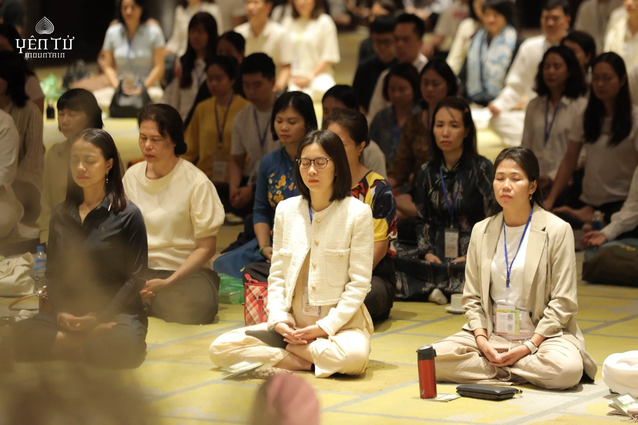 Group of people sitting on the floor, participating in a meditation or prayer session with closed eyes, in a large indoor conference or seminar setting.