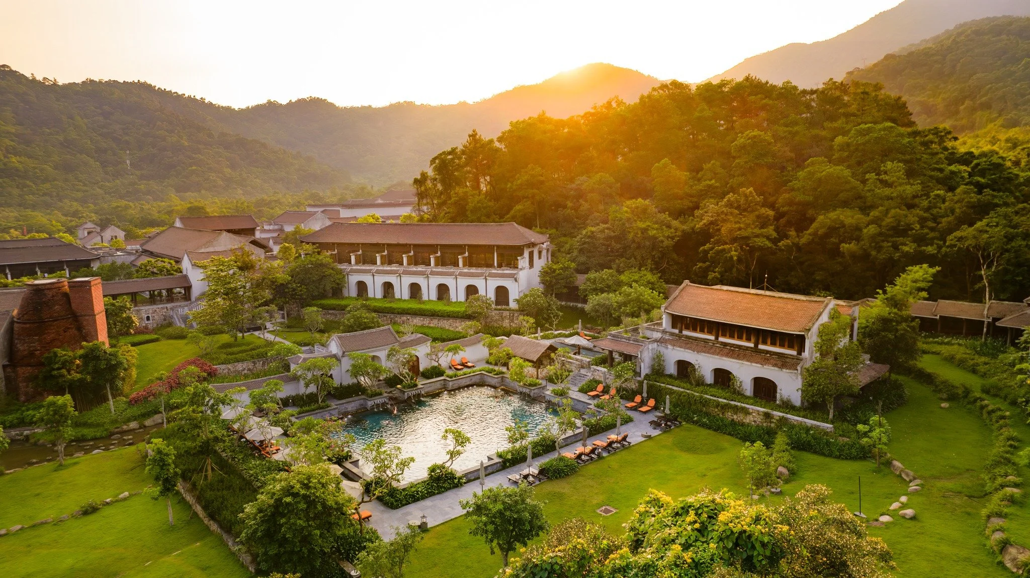 A scenic view of a resort with a swimming pool, surrounded by lush green gardens, multiple buildings with tiled roofs, and a forested mountain range in the background during sunset.