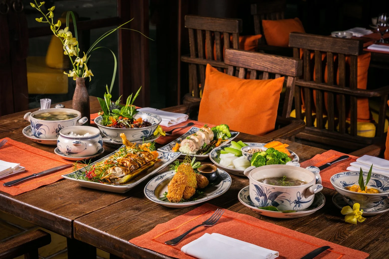 A wooden dining table set with various dishes and beverages, decorated with a tall vase of yellow orchids, surrounded by orange cushioned chairs, in a cozy restaurant setting.