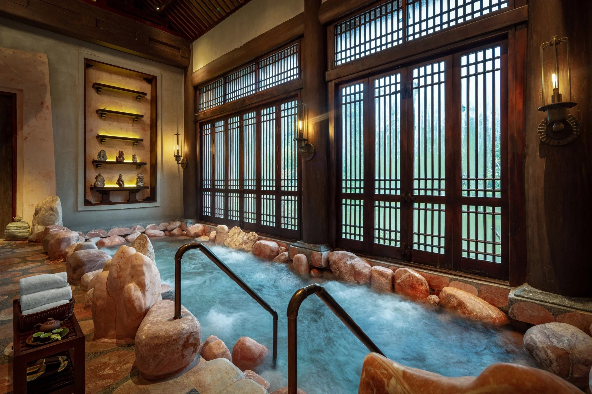 Indoor Japanese-style bath with large rock border, wooden lattice windows, and soft lighting.