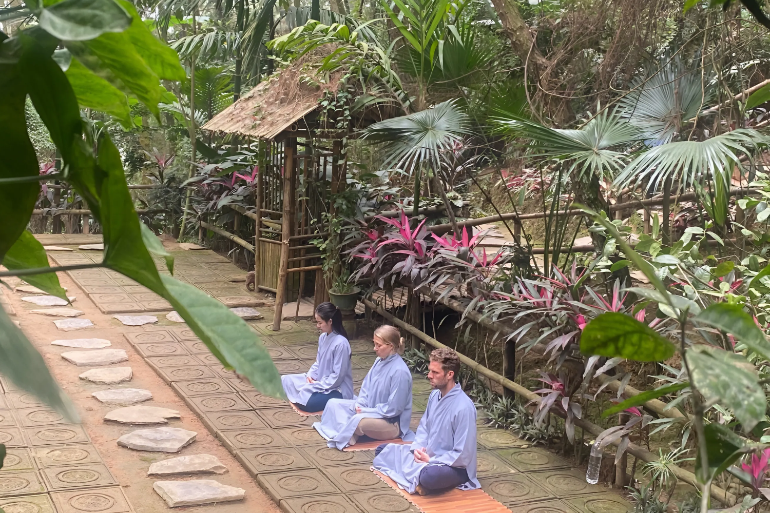 Three people practicing meditation in prayer position, kneeling on mats outdoors surrounded by lush tropical plants.