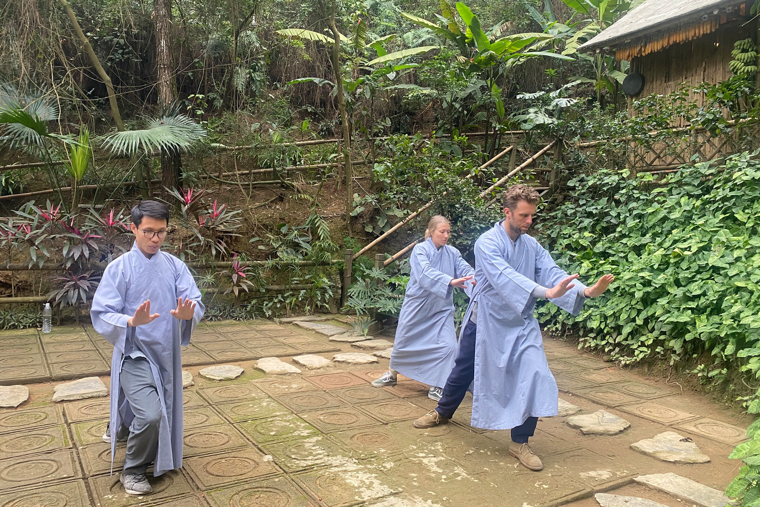 Three people practicing Tai Chi outside on a paved area surrounded by lush green plants in a garden or forest setting.
