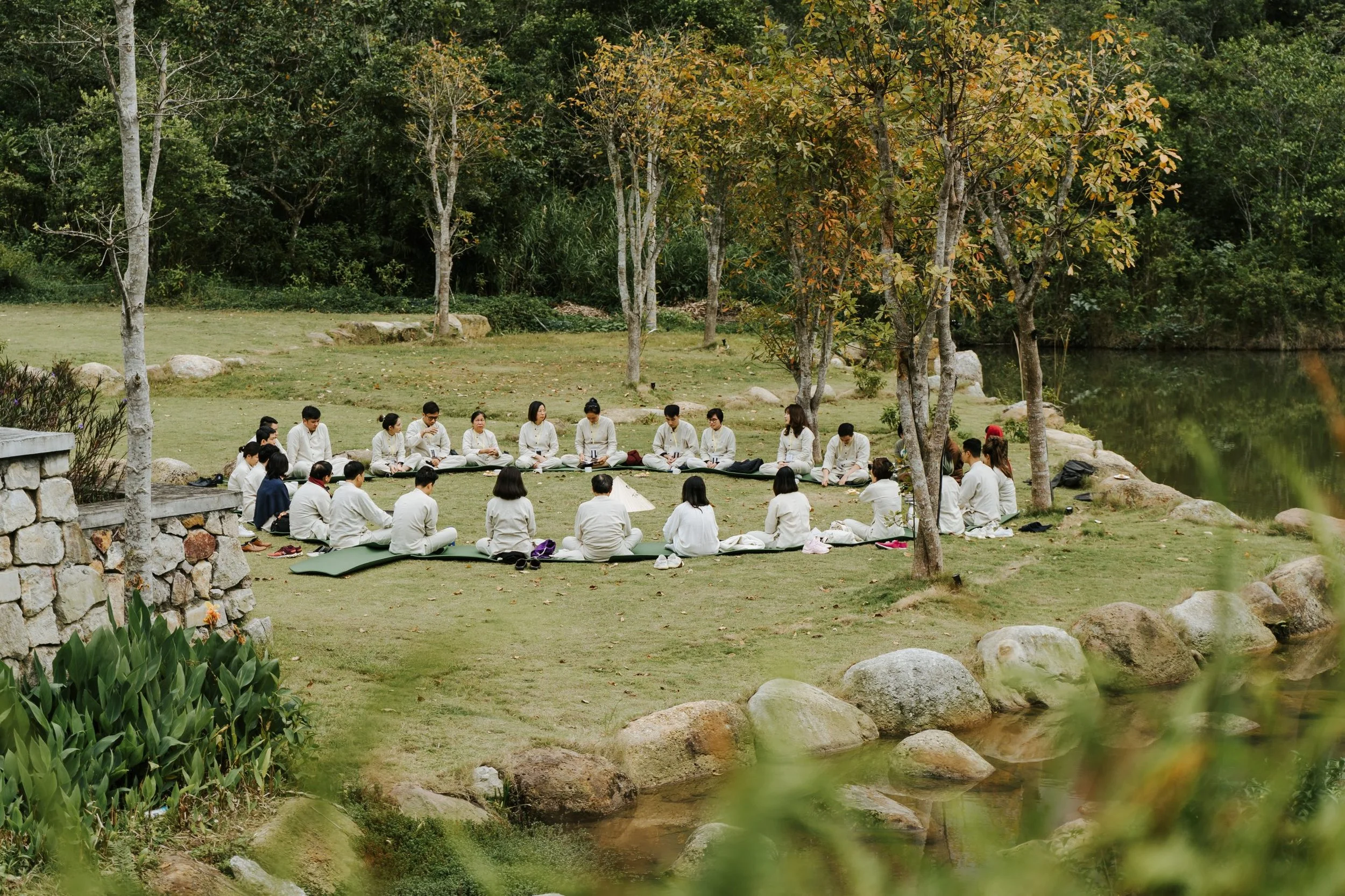 Group of people practicing yoga outdoors near a lake, sitting in a circle on mats surrounded by trees and rocks.