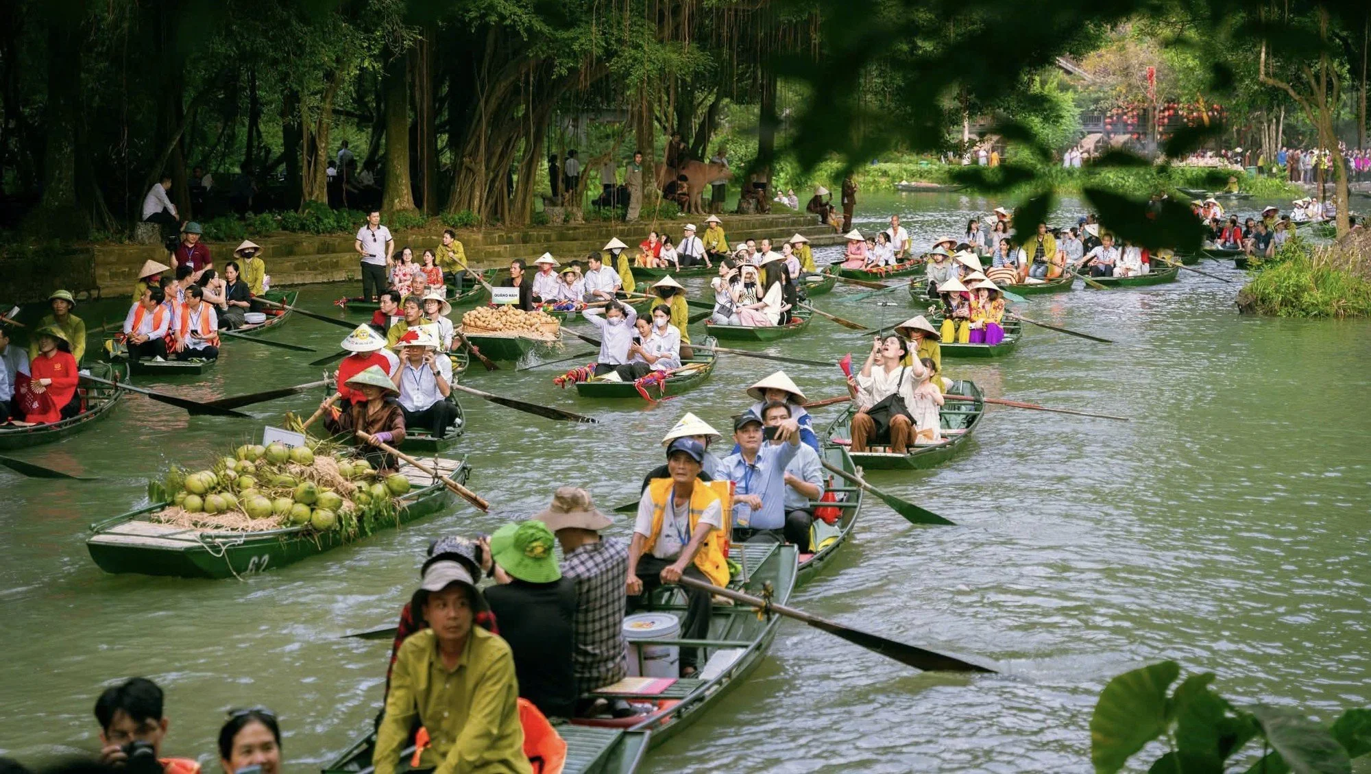 Multiple small boats filled with people, some wearing traditional conical hats, floating on a river surrounded by lush trees, with vendors selling fruits and tourists enjoying the scenery.