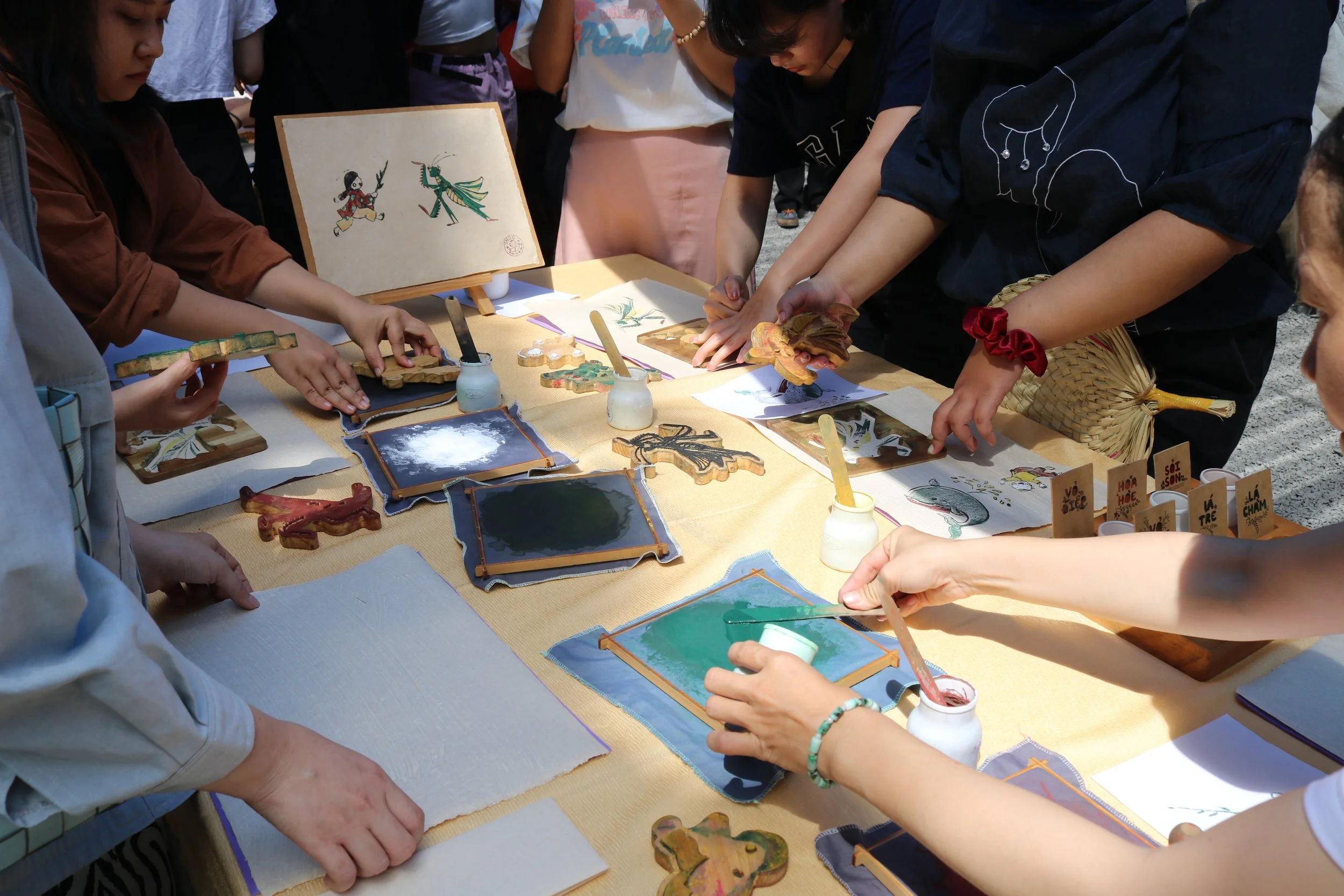 People gathered around a table engaging in traditional Japanese craft activity, using brushes and ink on glass plates, with decorative wooden cutouts and drawings of Japanese folklore on display.