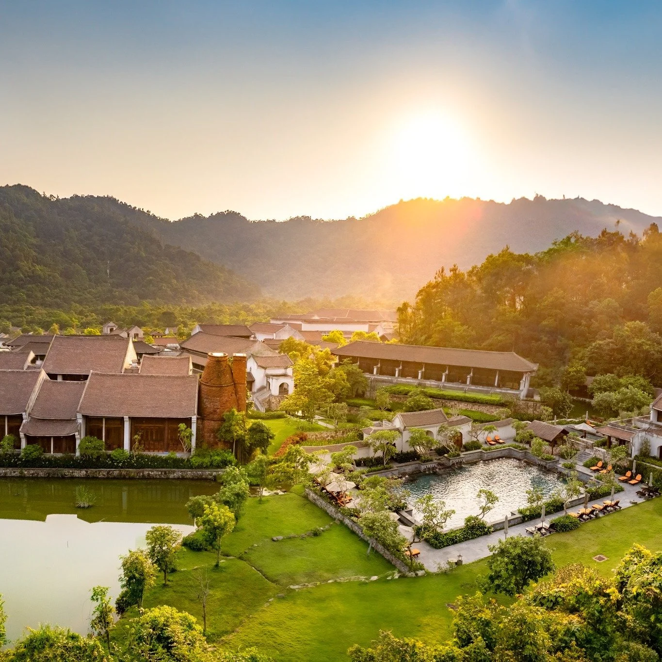 A scenic view of a resort with traditional-style buildings, a swimming pool, and greenery, set against a backdrop of forested mountains at sunset.