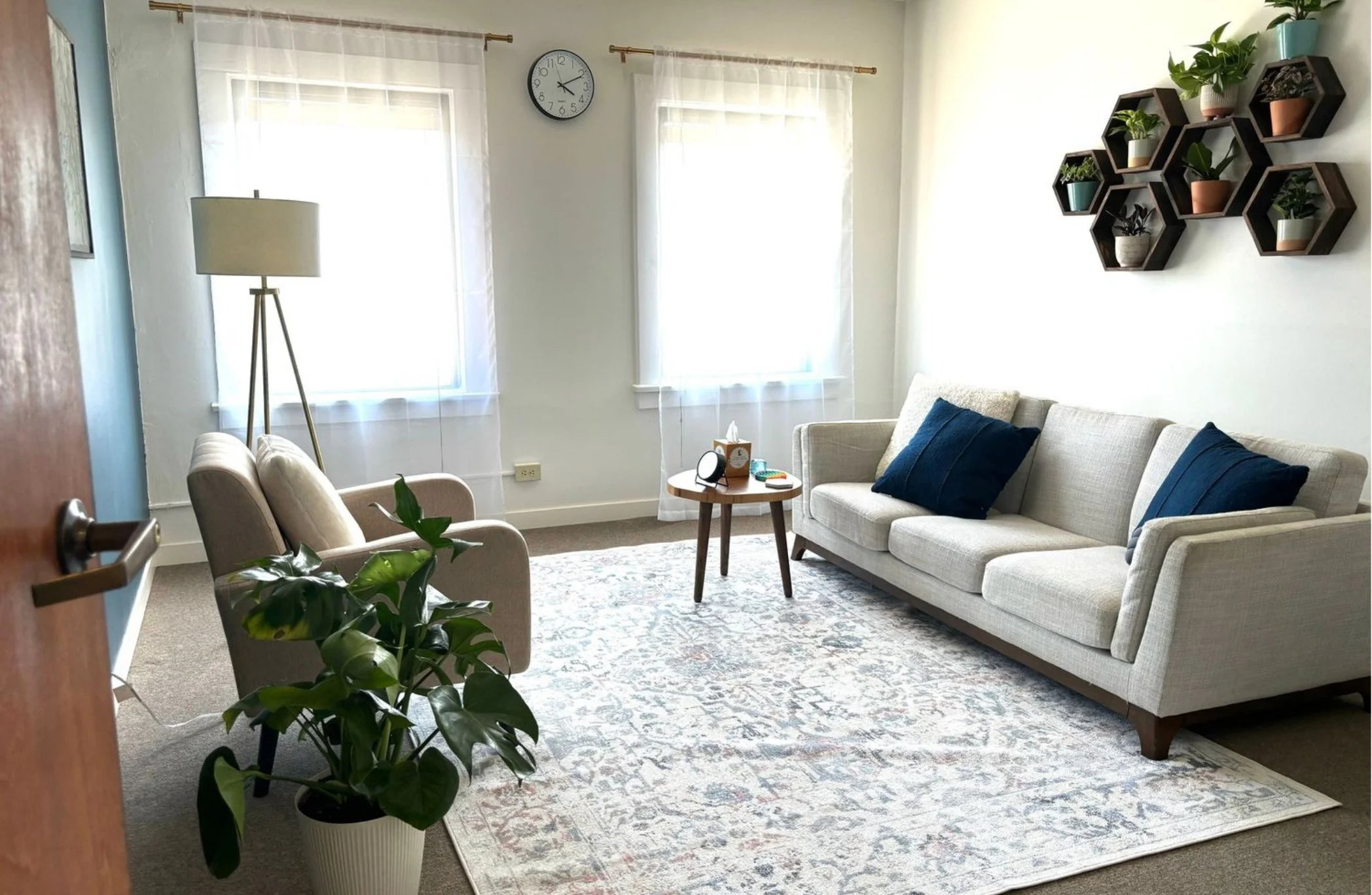 A therapy office in Alhambra with a light-colored sofa, two dark blue pillows, beige armchair, potted plant, floor lamp, wall clock, three windows with white curtains.