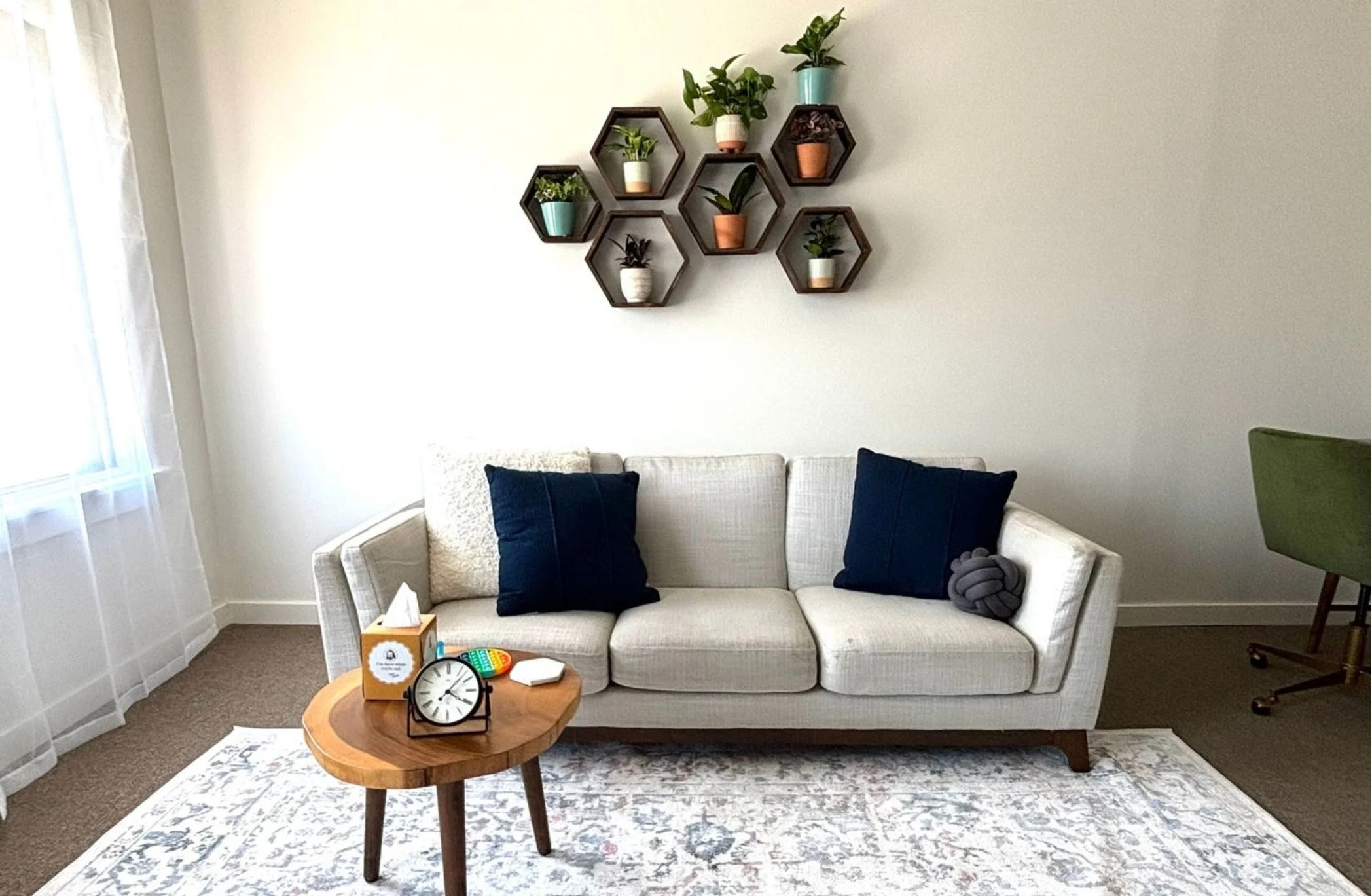 Therapy office in Alhambra with a white sofa, blue pillows, a wooden coffee table with a clock, tissues, and decorations, and wall-mounted hexagonal shelves with potted plants.