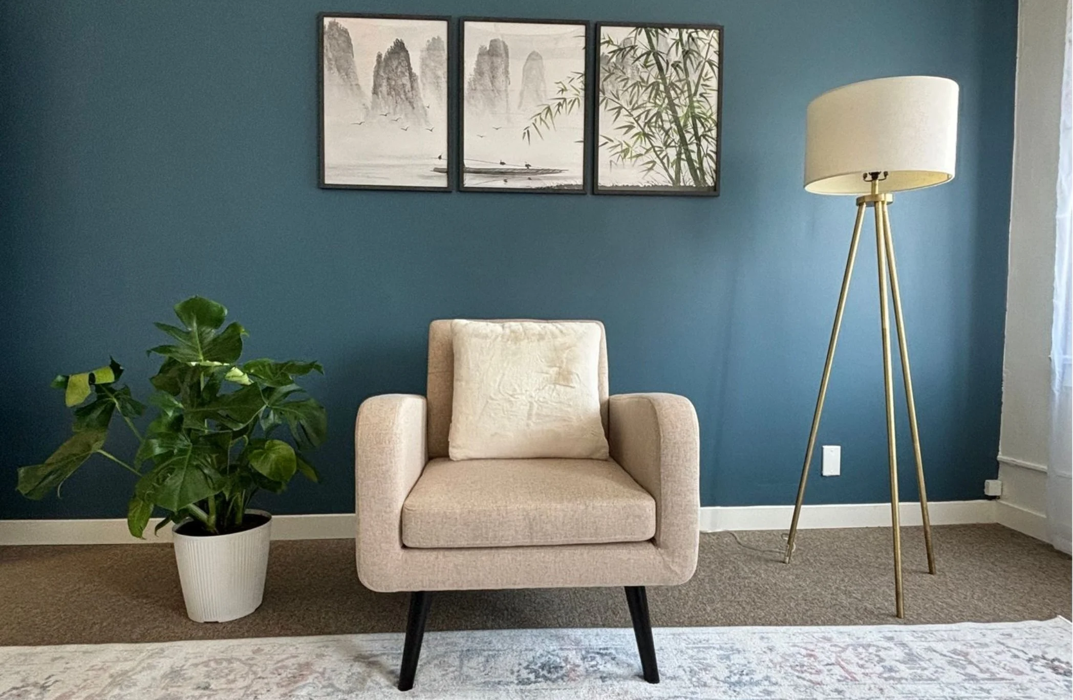 A cozy corner of Jorgie Wu Therapy office in Alhambra, CA with a beige armchair, a large potted plant, a tall floor lamp with a white shade, a triptych wall art with landscape and bamboo images, set against a teal wall.